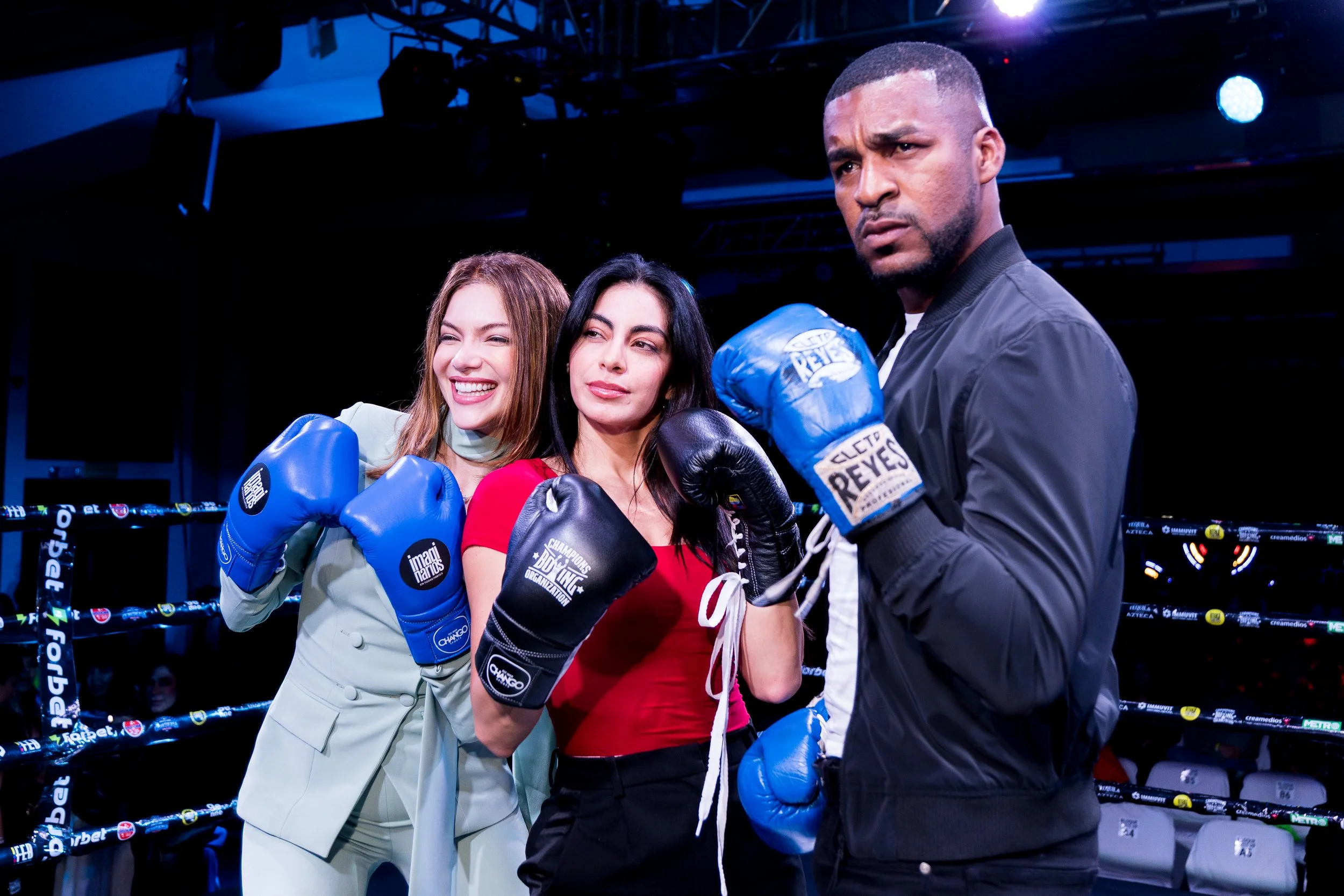 Three women with boxing gloves and a man with boxing gloves in a boxing ring. The women are smiling, and the man appears serious. The women are dressed in business attire, and the man is dressed in casual clothing. The background shows stage lighting