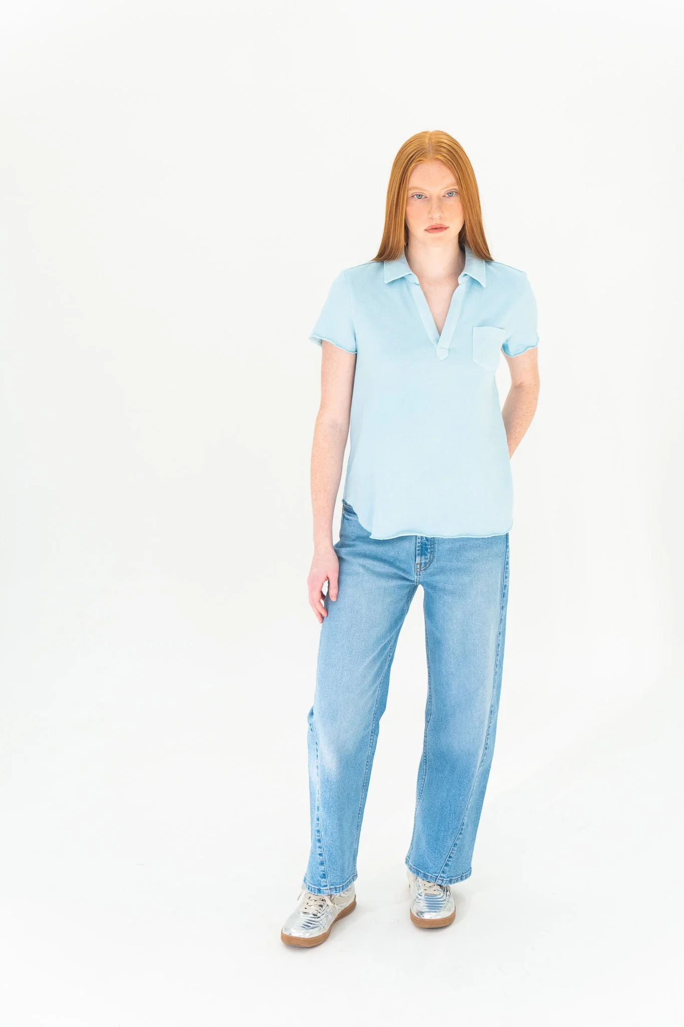 A young woman with long red hair wearing a light blue shirt, blue jeans, and white sneakers standing against a plain white background.
