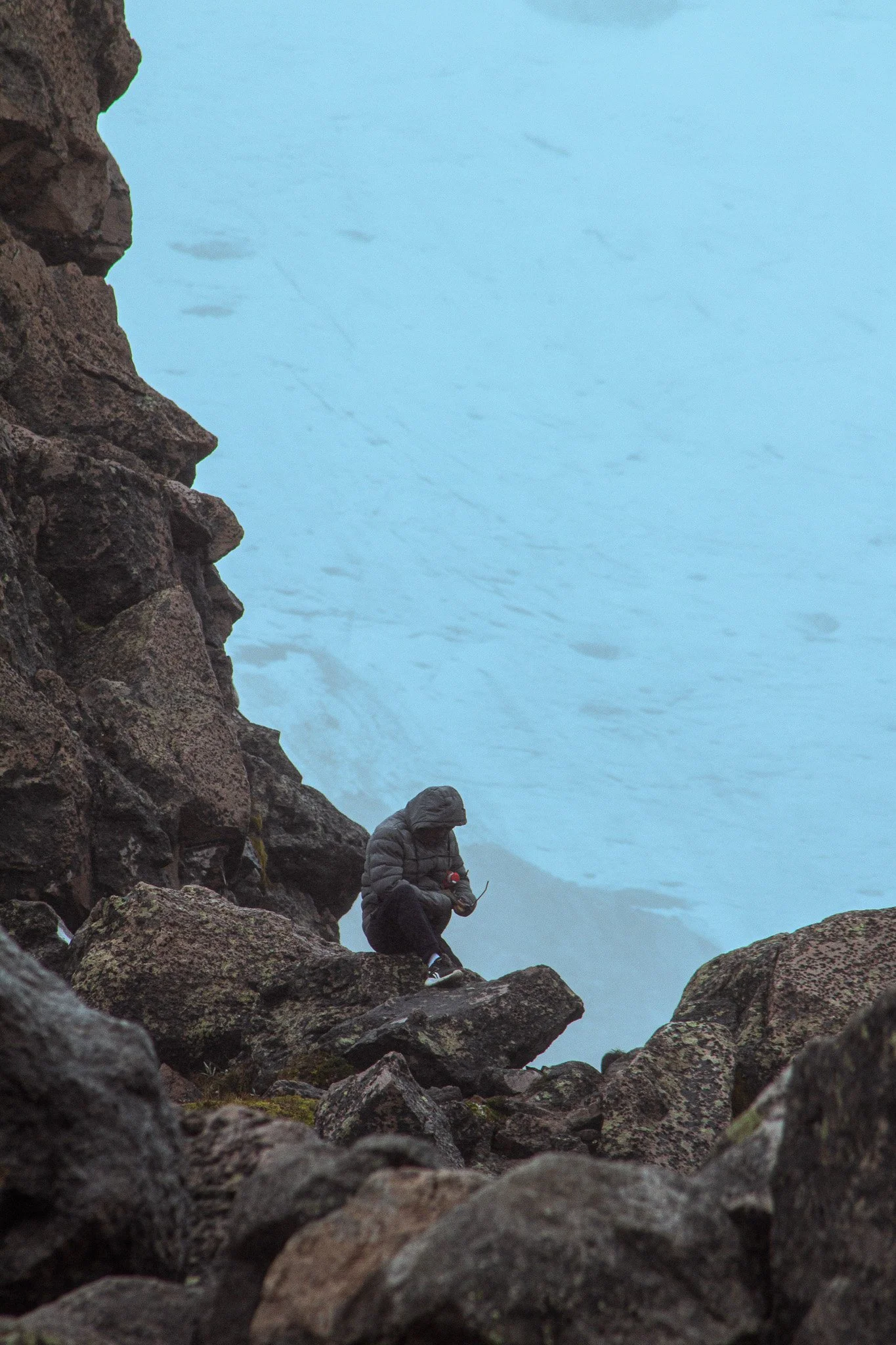Person in a gray jacket sitting on rocks near a body of water, looking at a device, surrounded by large rocks.