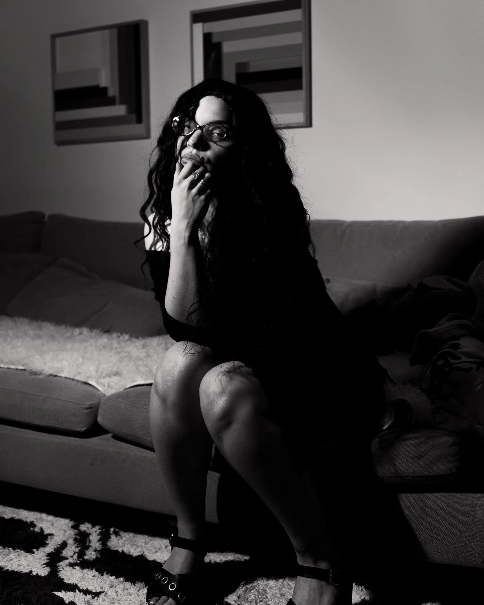 A black and white photo of a woman sitting on a couch, leaning on her knees with her hand near her face, wearing glasses and textured clothing. There are abstract framed artworks on the wall behind her, and a fluffy rug and cushions on the floor.