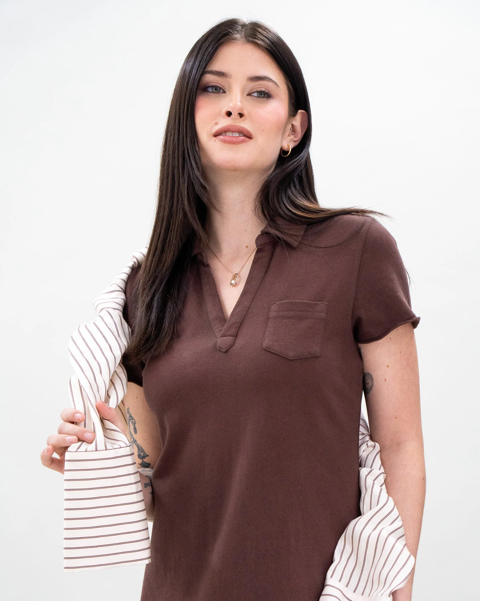 A woman with long dark hair and tattoos on her left arm is wearing a brown shirt and a striped blazer draped over her left shoulder, standing against a plain white background.