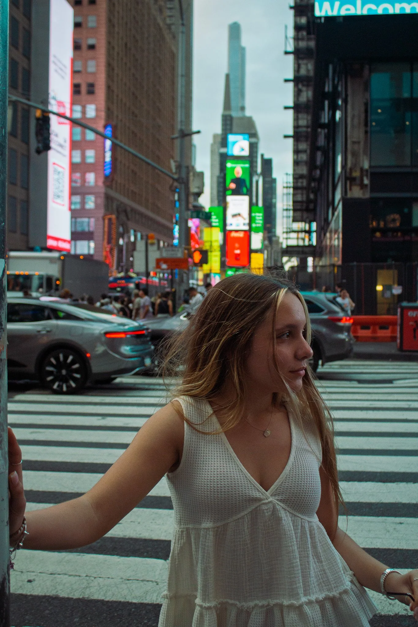 A young woman with long, wavy, blonde hair stands near a crosswalk on a busy city street, with tall buildings and bright digital billboards in the background.