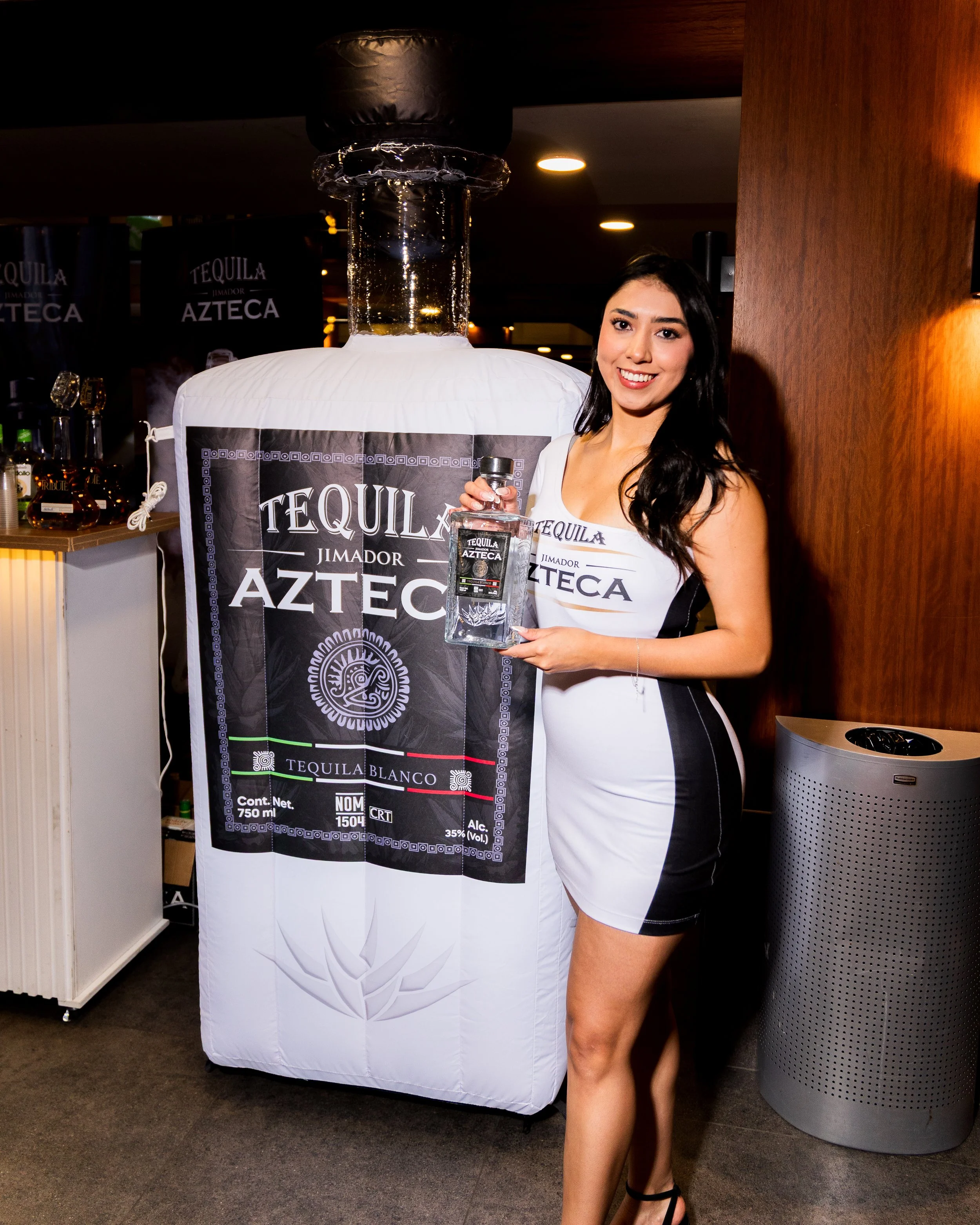 A woman with long dark hair smiling and holding a bottle of tequila. She stands next to a large banner with tequila branding, including a graphic measuring alcohol percentage and volume. The setting appears to be an indoor event or exhibition.