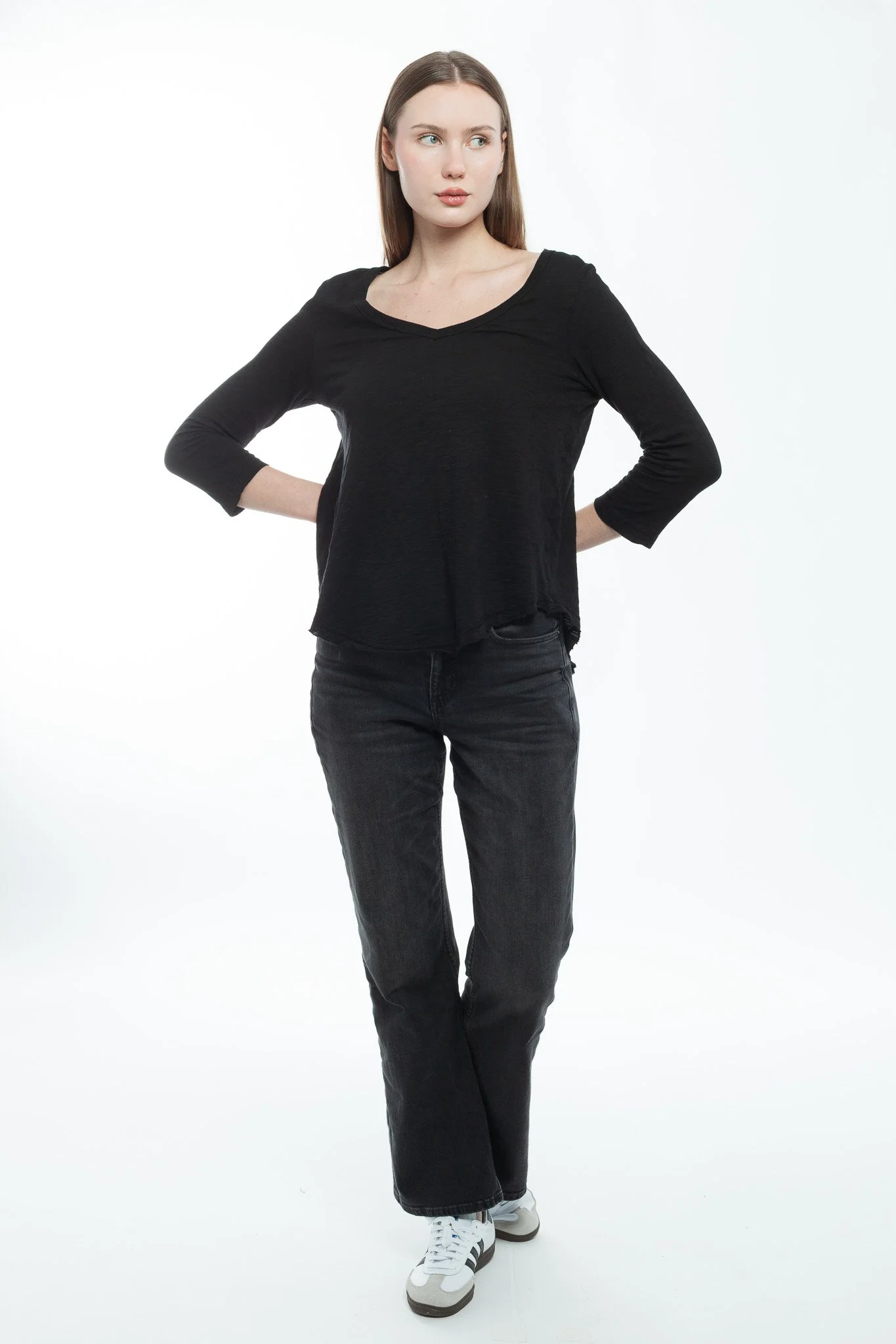 A young woman with long brown hair, wearing a black long-sleeve shirt, black jeans, and white sneakers, standing against a plain white background with her hands on her hips.