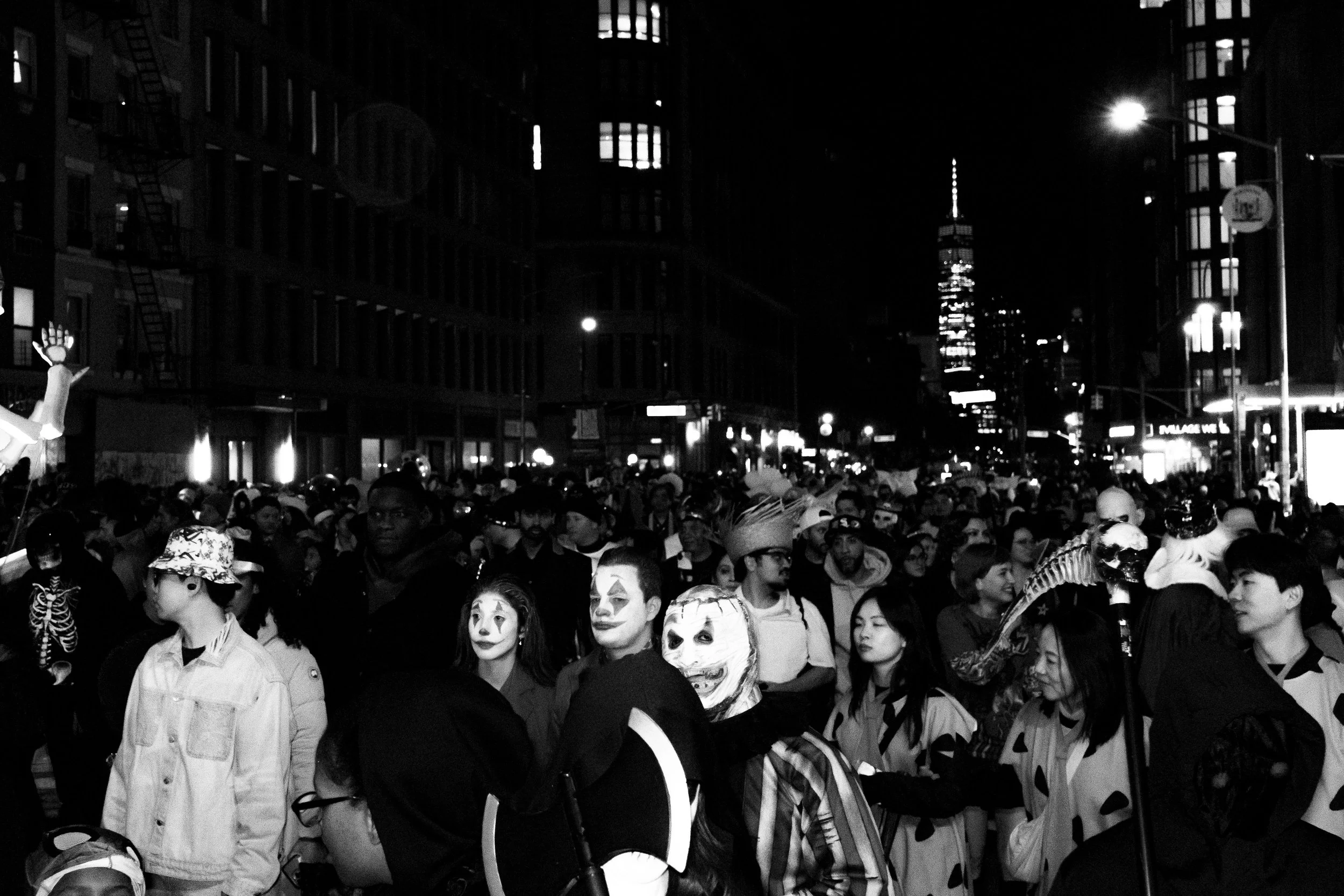 A large crowd of people dressed in costumes and masks gathered on a city street at night, with tall buildings and city lights in the background.