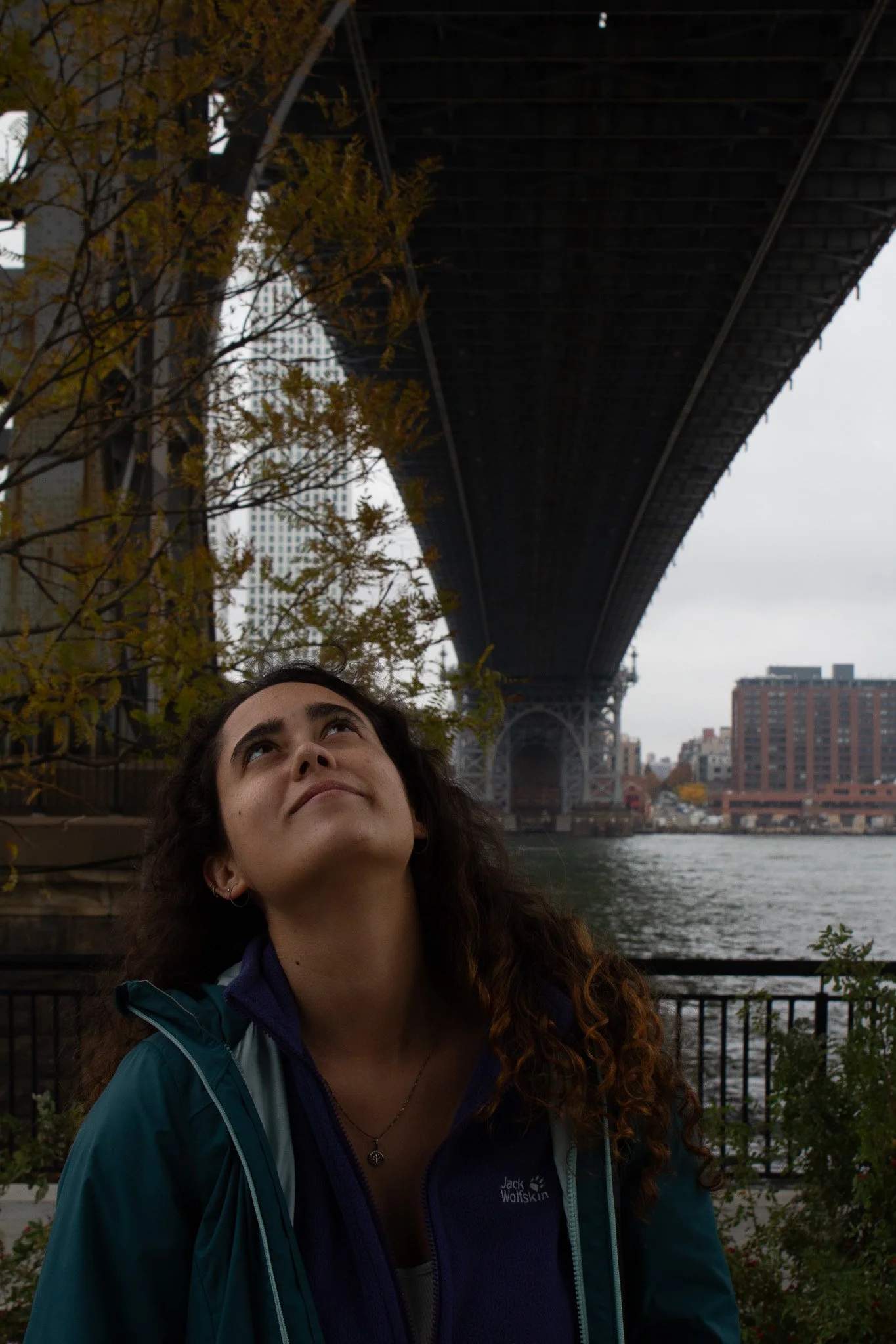 A woman with curly hair looking up at a large bridge overhead near a river in an urban area with buildings and trees around.