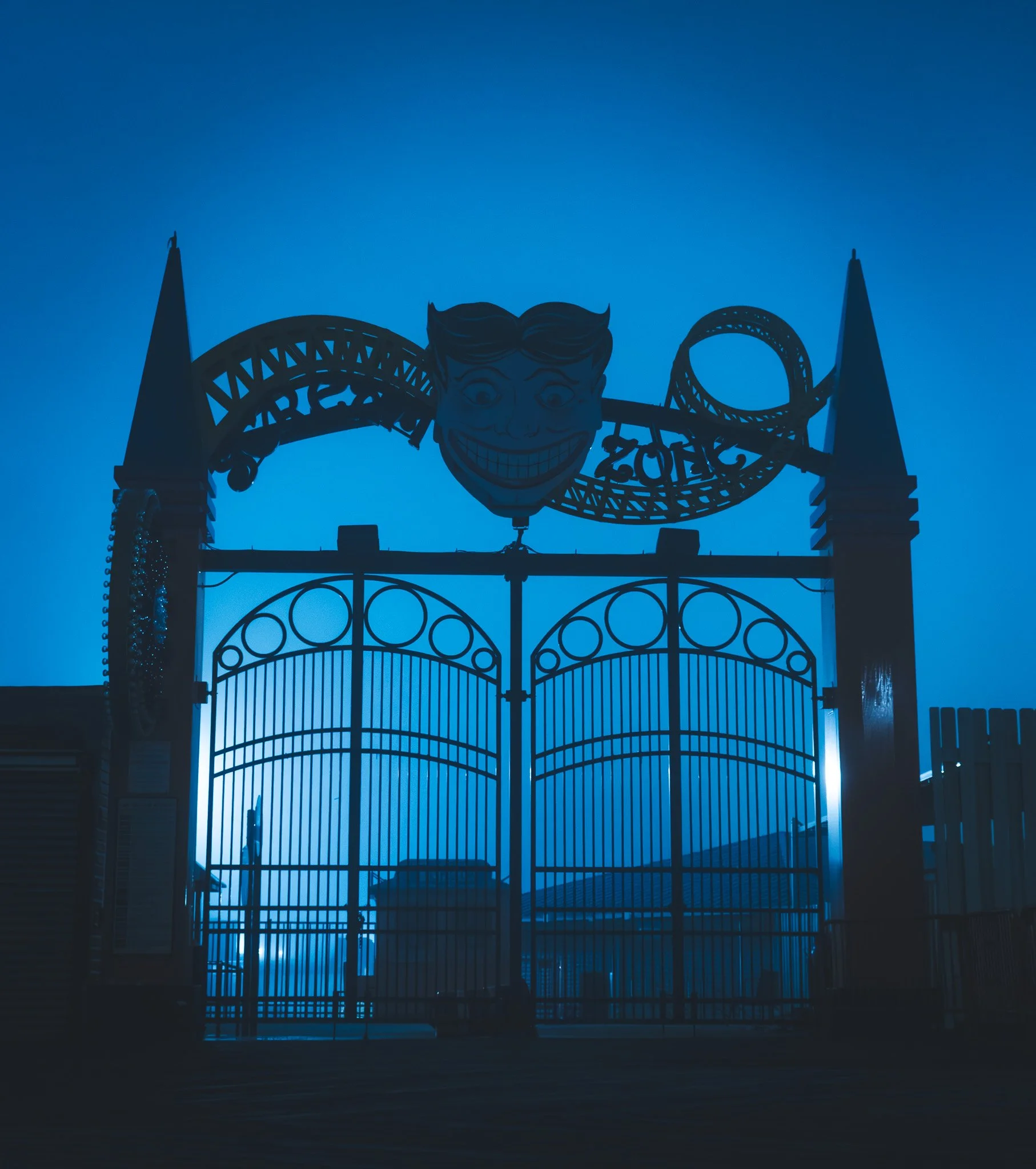 Silhouette of a roller coaster and a clown face logo against a blue twilight sky, possibly at an amusement park entrance.