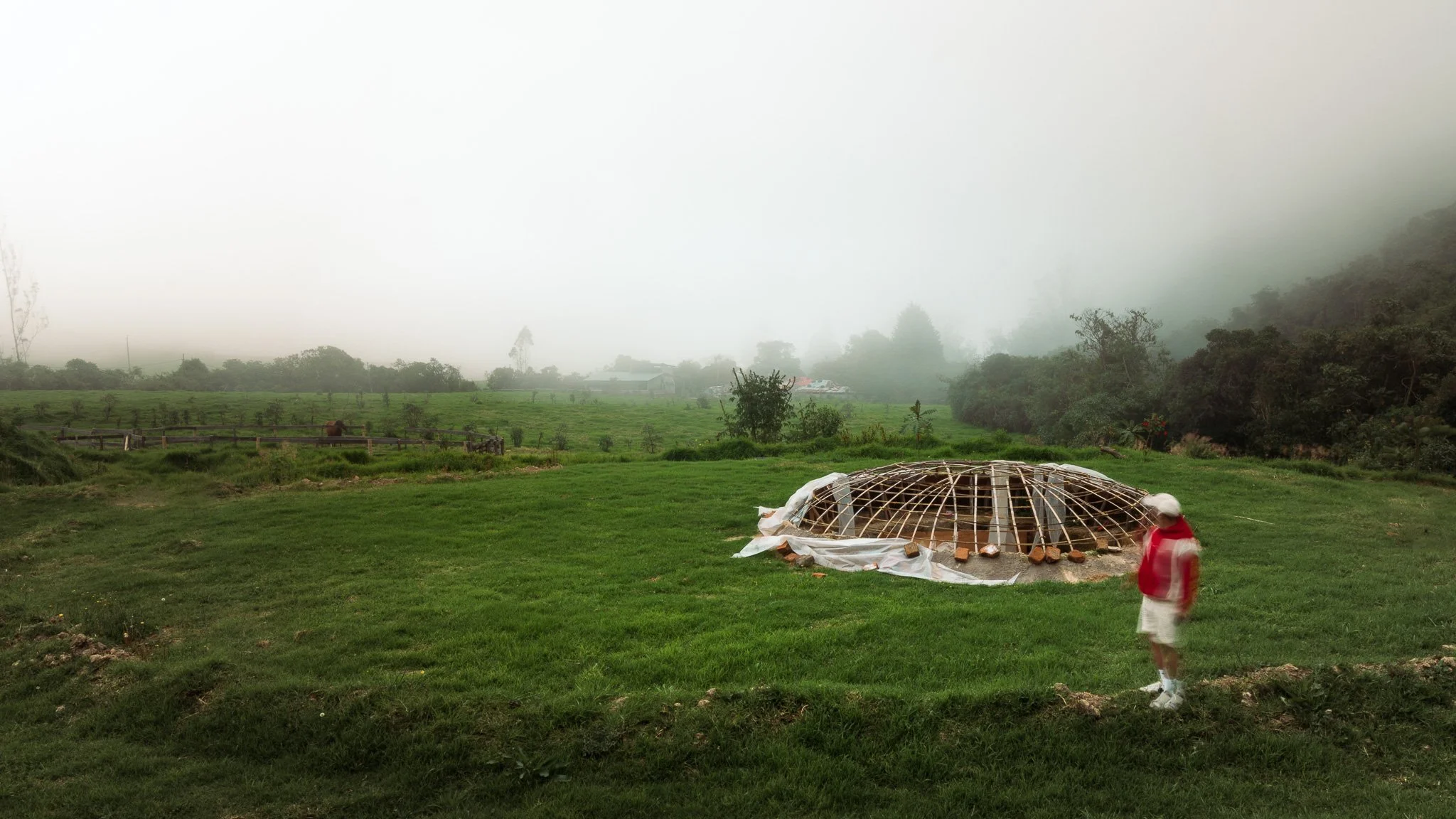 A person in a red jacket and white pants walking past a large framework of wood and plastic sheeting on a grassy field, with trees, a fence, and misty hills in the background.