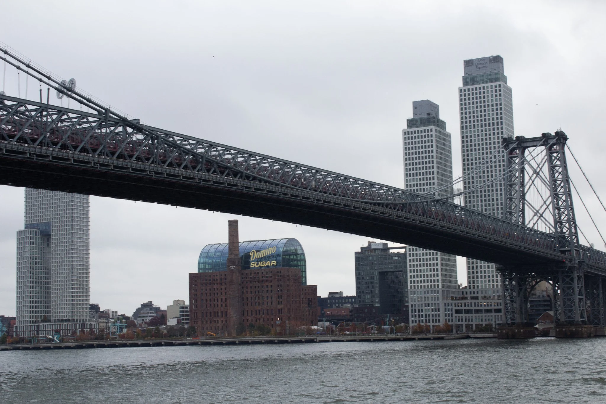 A city skyline with a bridge over a body of water, tall modern buildings including the Domino Sugar building with a large sign, and an overcast sky.