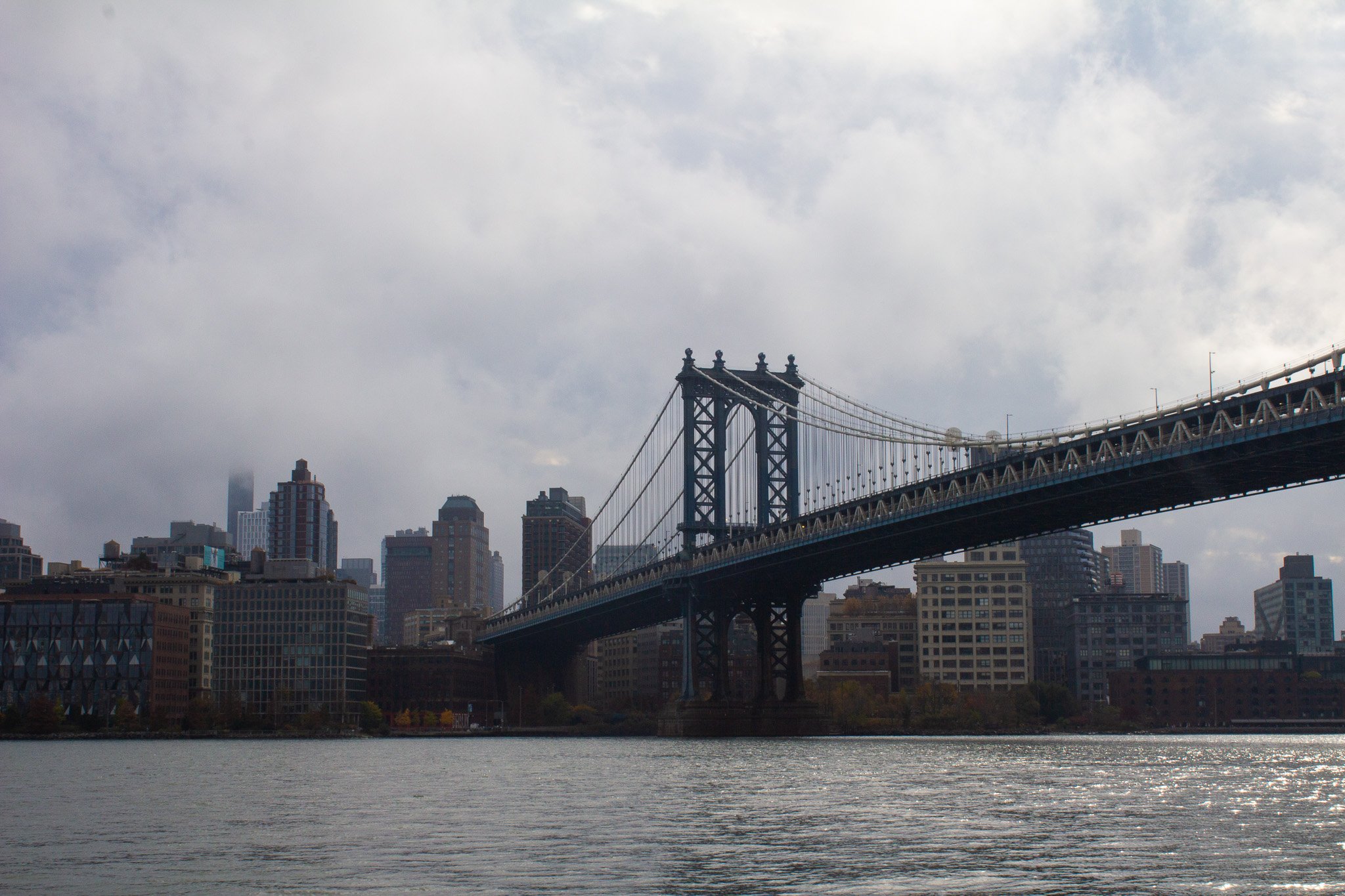 View of a suspension bridge over a river with a city skyline, including skyscrapers, in the background on a cloudy day.