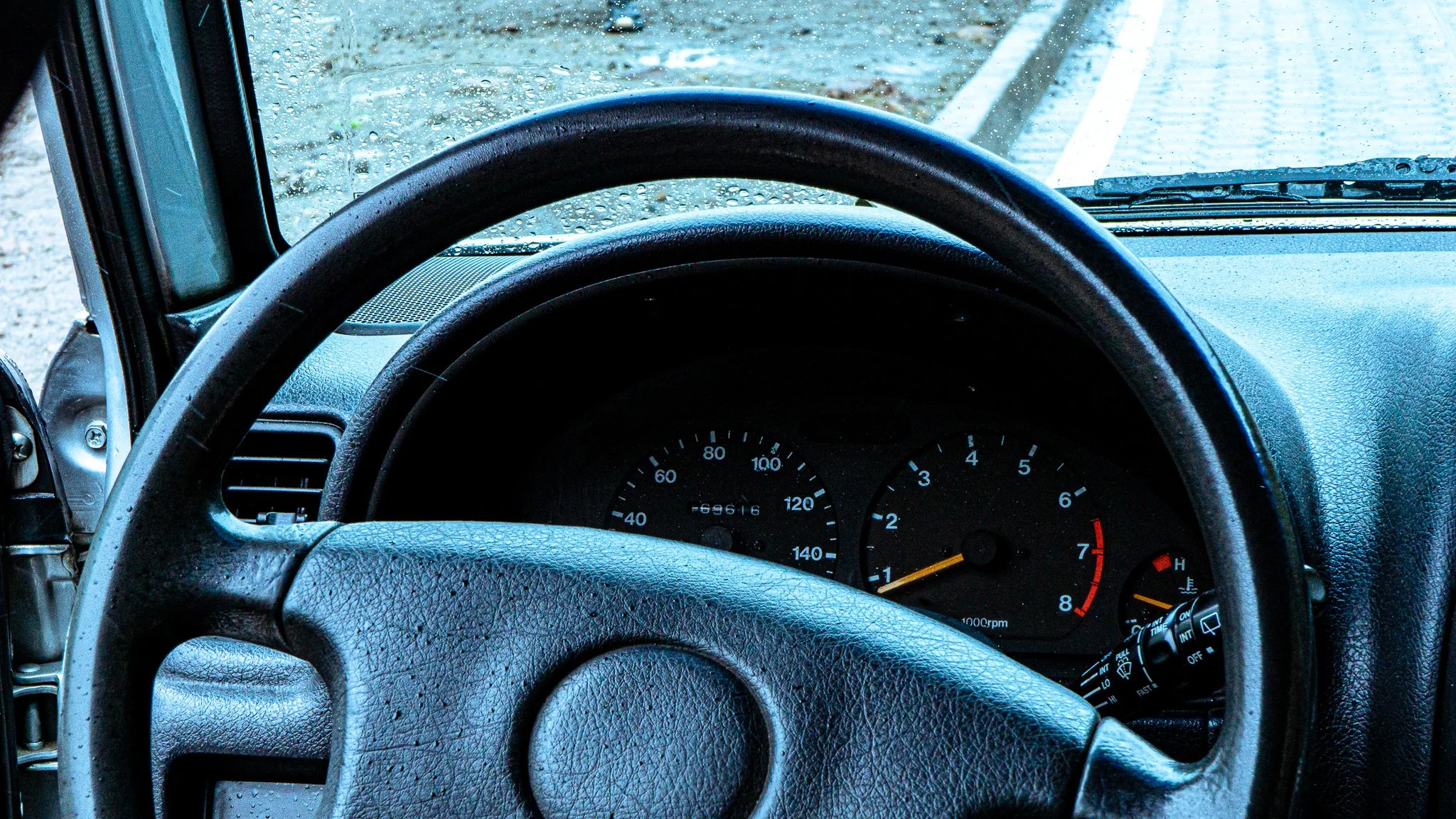 Inside the cab of a vehicle showing a black steering wheel with water droplets on it, dashboard with speedometer, and rain outside the windshield.