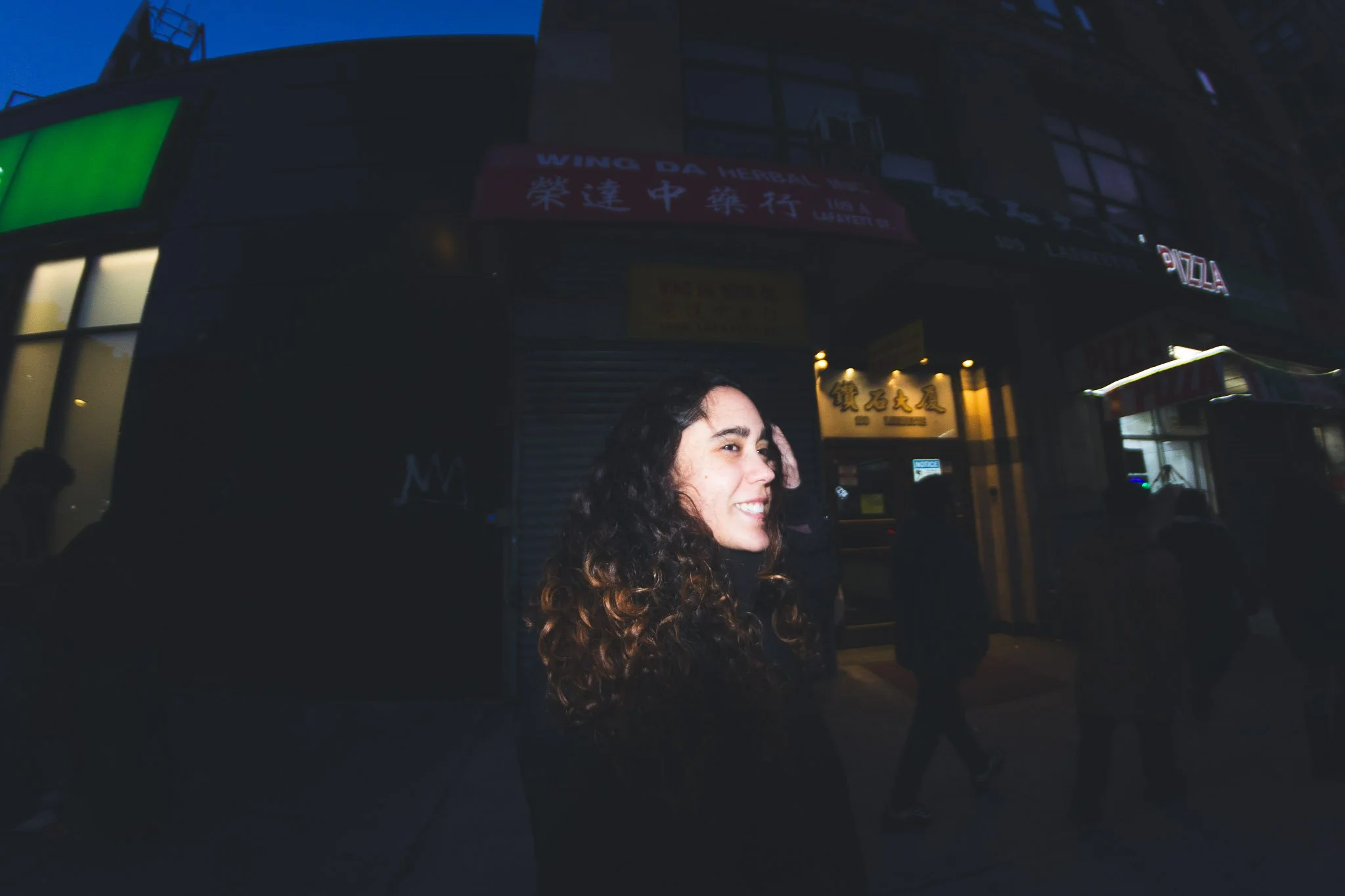 A woman with curly hair smiling and walking on a city street at night, with illuminated storefronts and signs in the background.