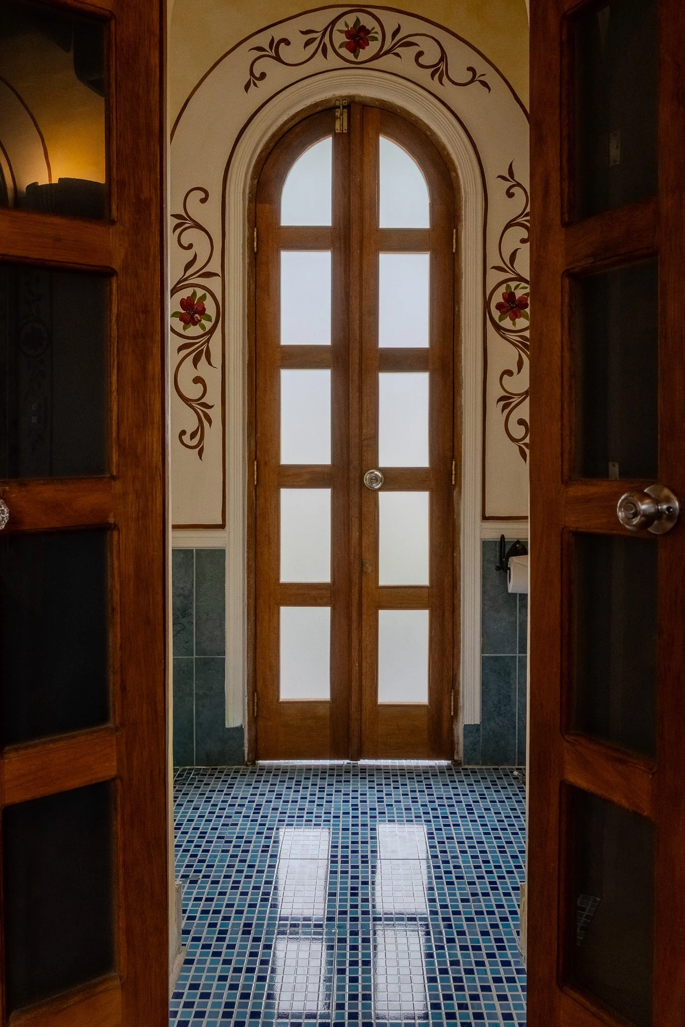 A view through wooden door frames showing a wooden door with frosted glass panels, a tile floor with a pattern of blue, white, and black tiles, and a partly visible wall with floral decorations.