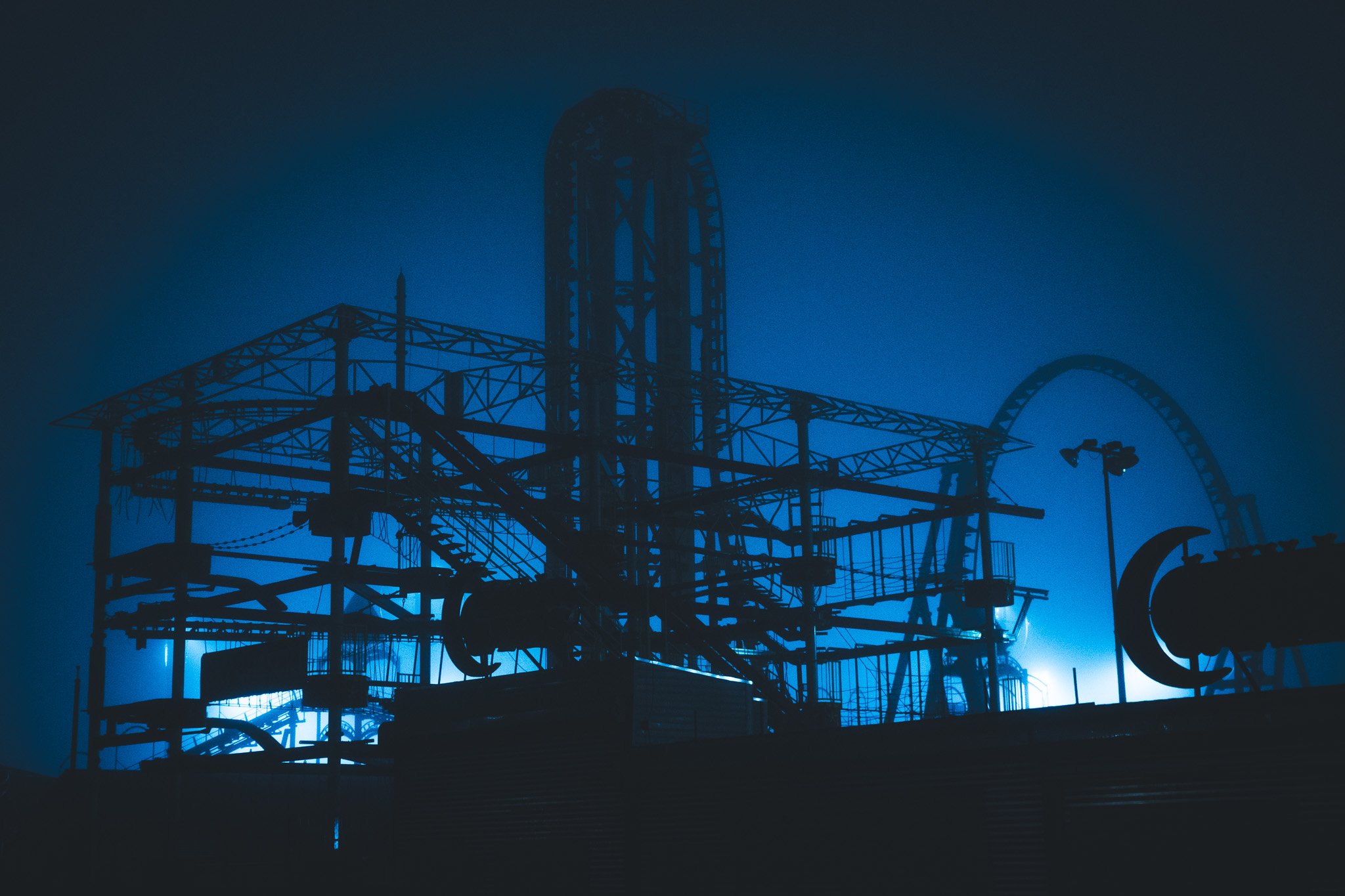 Silhouette of an industrial roller coaster or ride against a dark blue sky, with intricate metal framework and a faint light illuminating part of the structure.