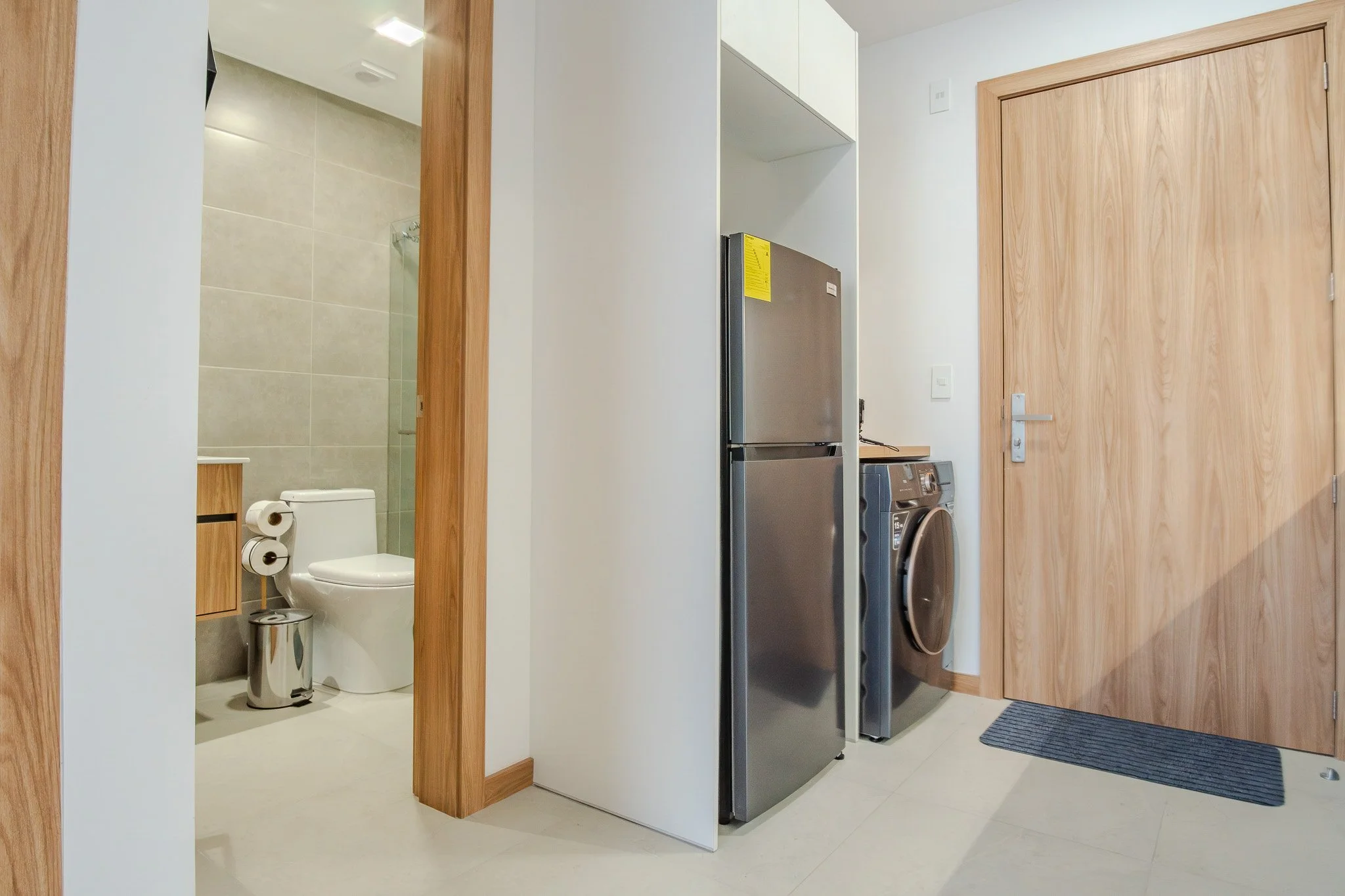 Part of a modern apartment or house interior showing a bathroom with a toilet and a separate laundry area with a washing machine, a refrigerator, and a wooden door.