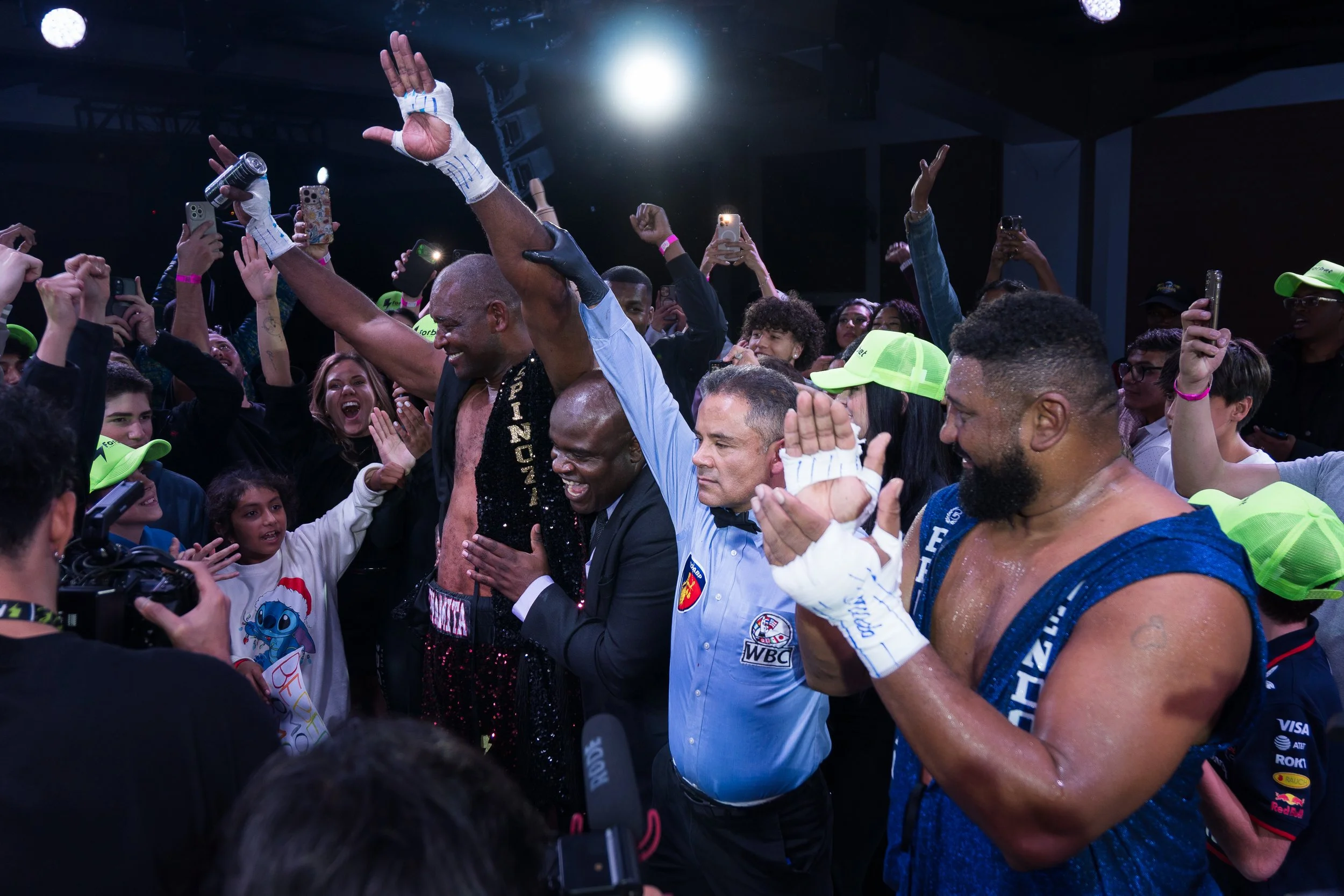 A group of people, including a shirtless boxer with hand wraps and a suit with the World Boxing Council (WBC) logo, are celebrating in a crowded indoor setting. The boxer appears to have just won a match, surrounded by fans taking photos and cheering