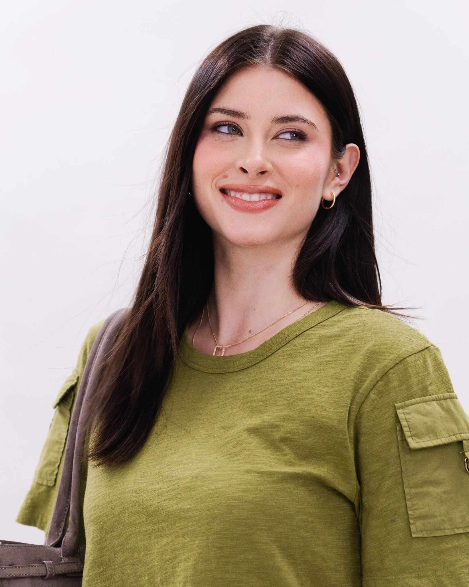Close-up of a woman with long dark hair wearing gold earrings and a necklace, looking to the side with a slight smile, dressed in a green top with rolled-up sleeves, against a plain white background.