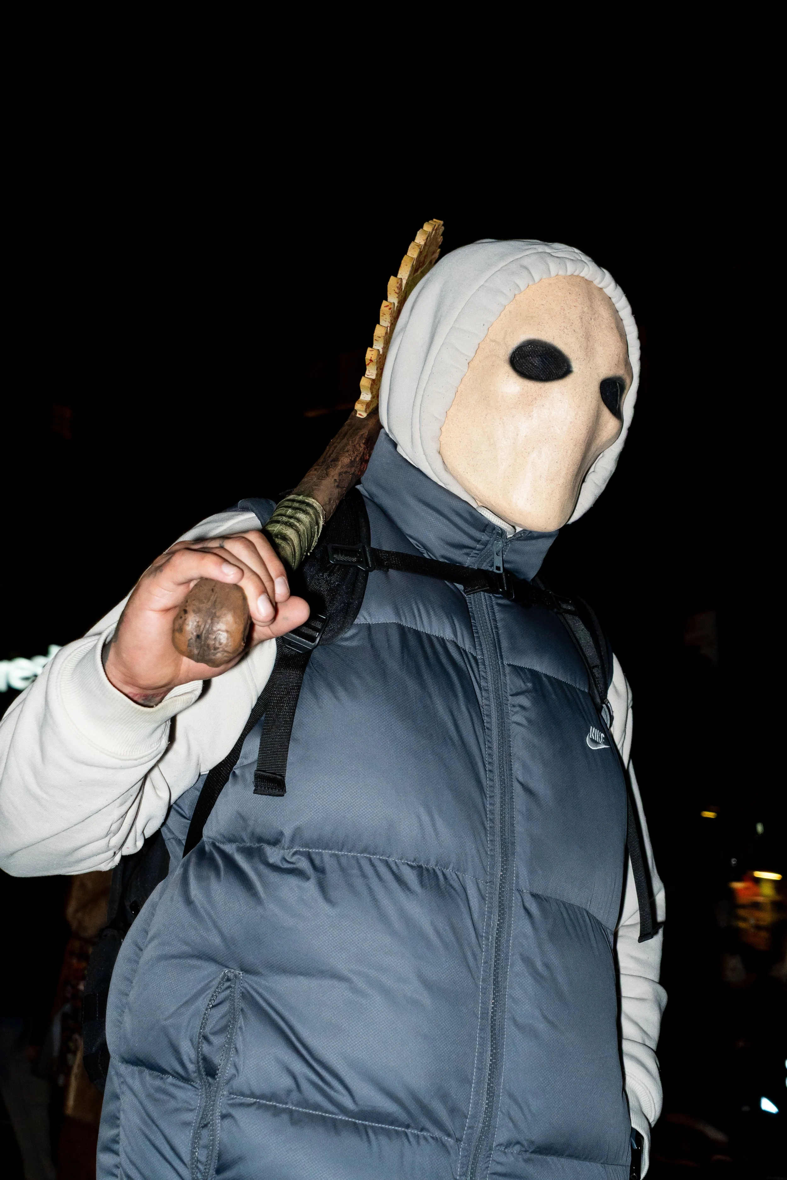 Person wearing a mask resembling a creepy face with black eye holes, dressed in a jacket, carrying a wooden stick over their shoulder at night.