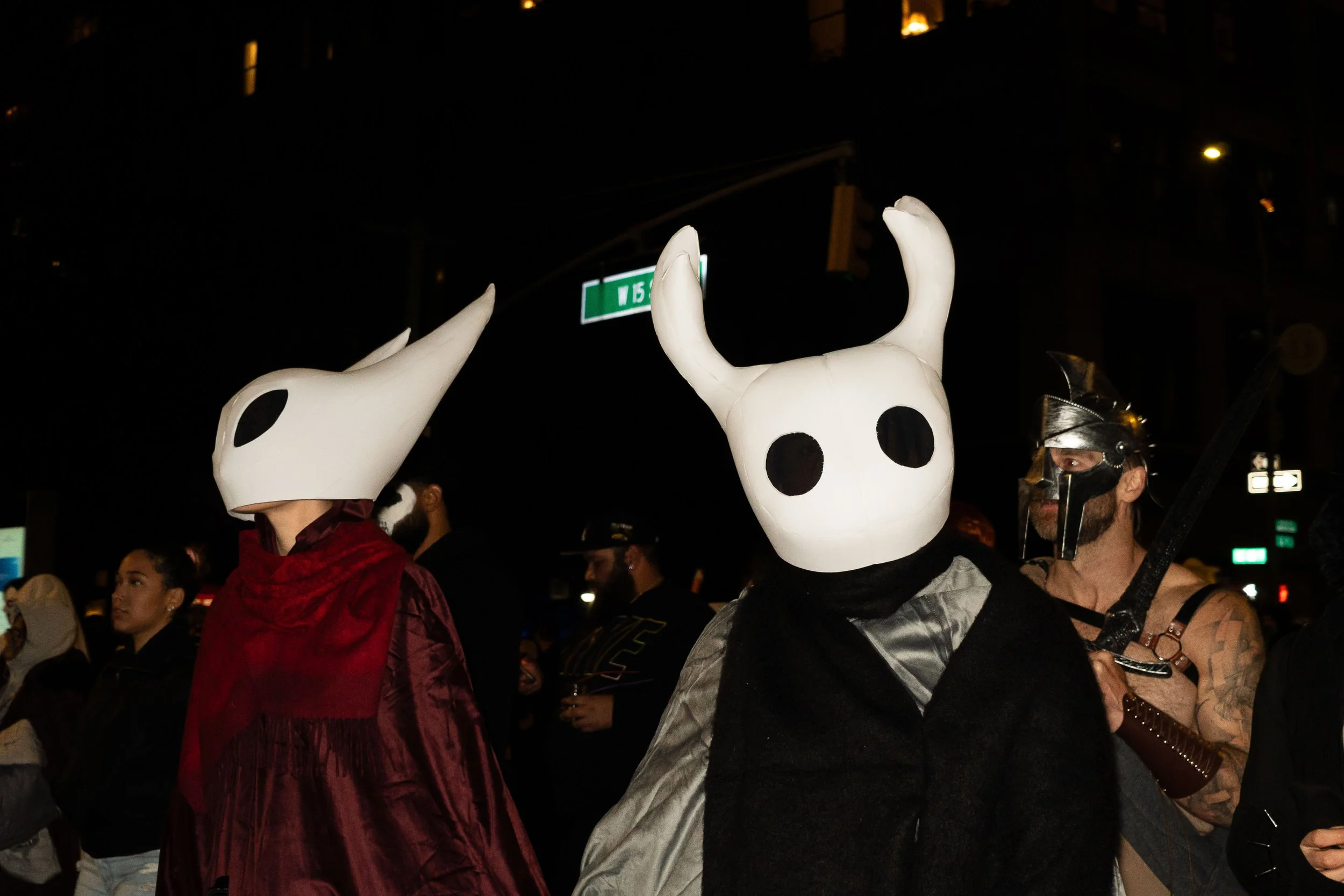 People dressed in costumes with masks, including one with a large white mask with horns and black eye holes, and another with a black mask and armor, at night.