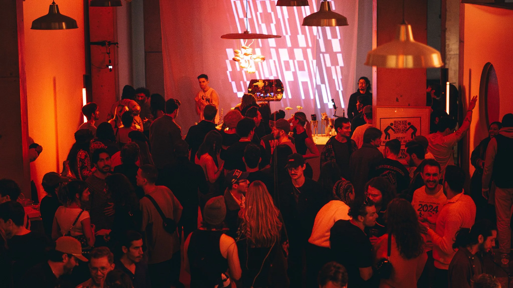 A crowded indoor event with many people standing and socializing under warm lighting, with a large screen or projection on the wall displaying geometric patterns.