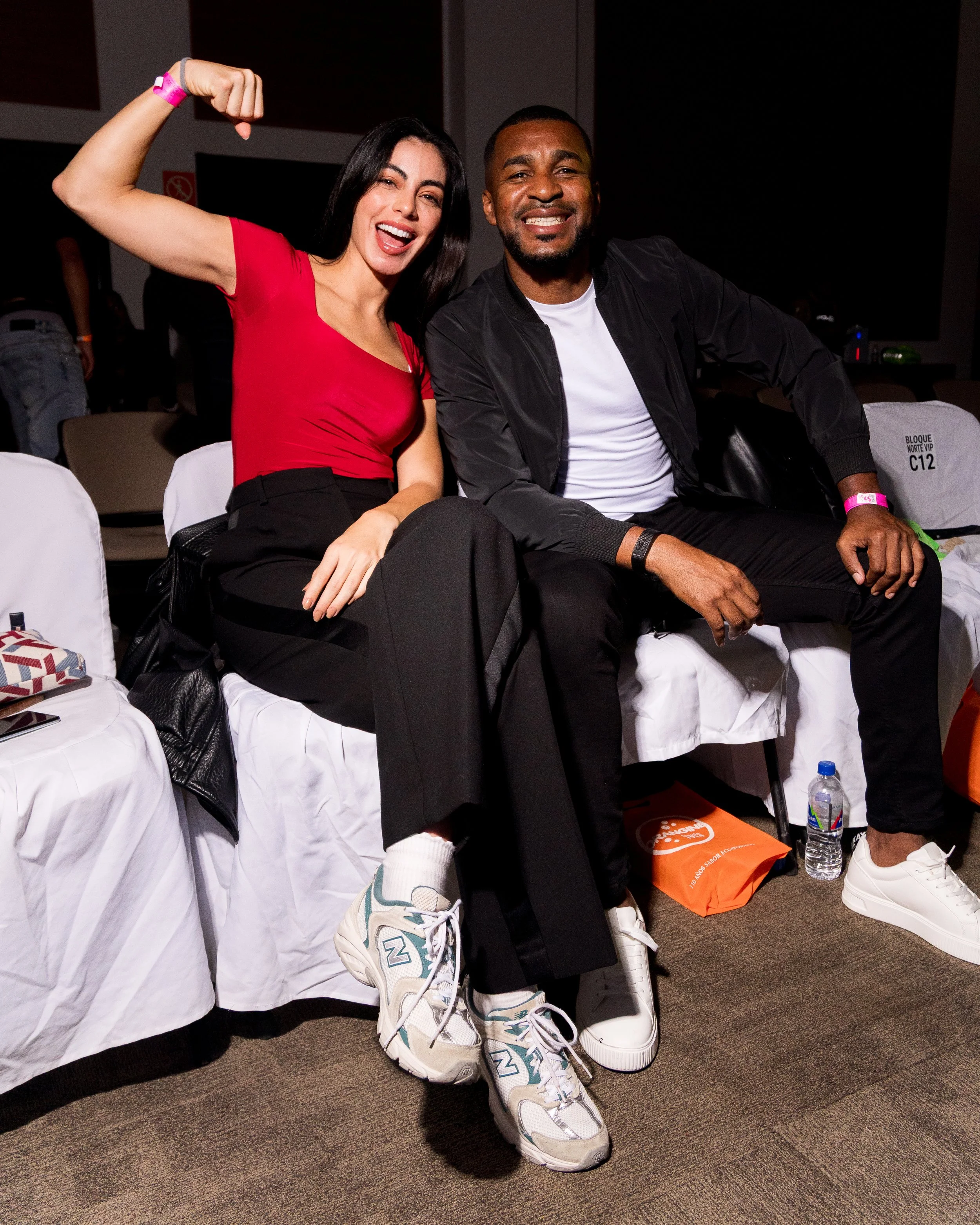 Two people sitting on chairs at an event, smiling at the camera. The woman on the left is flexing her bicep and wearing a red top, black pants, and white sneakers. The man on the right is wearing a black jacket, white T-shirt, black pants, and white 