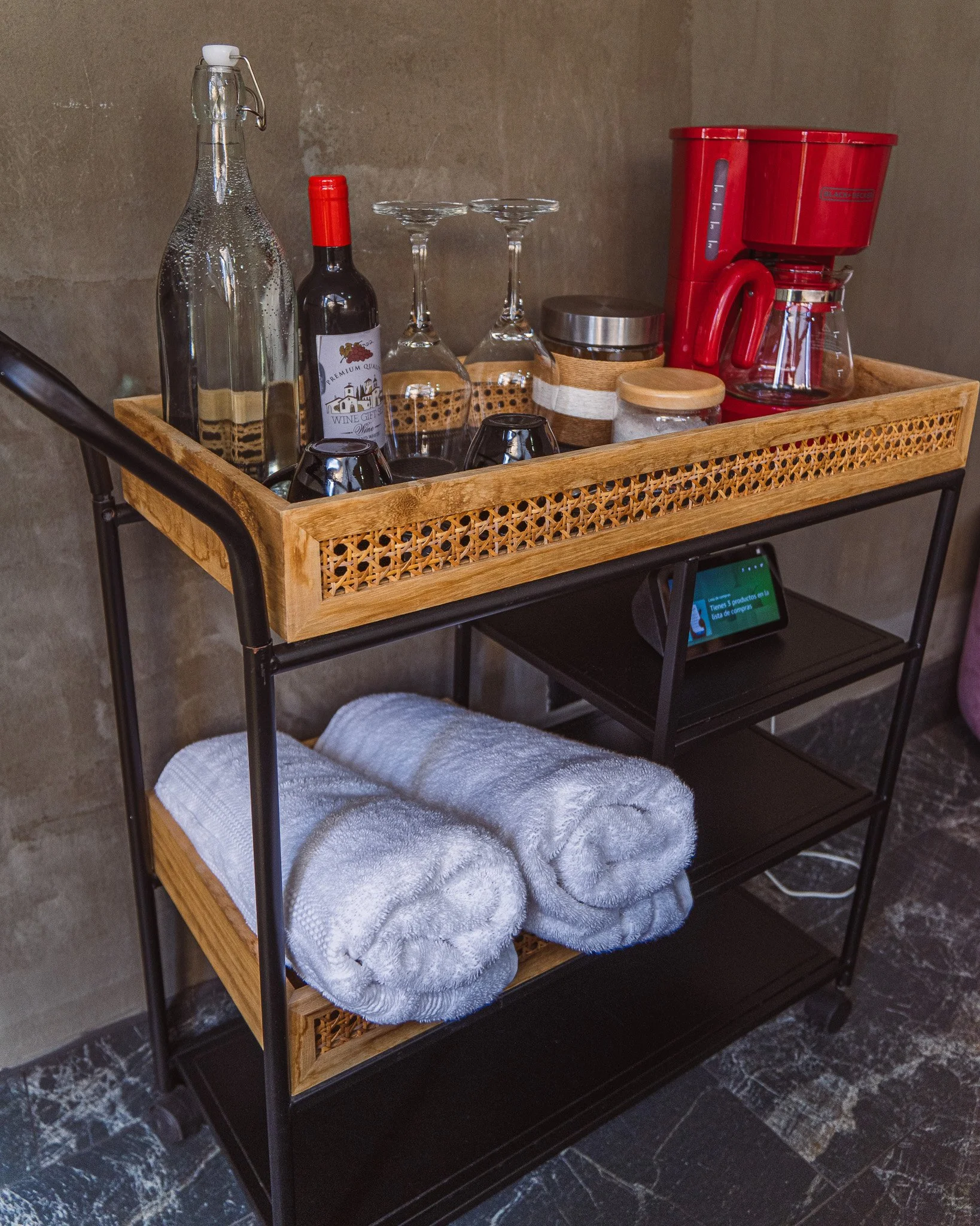 A wooden rolling cart with three shelves, containing a glass water bottle, a bottle of red wine, four glasses, a black container, a glass jar with a wooden lid, a red coffee maker, and a small appliance. Towel is draped over the lower shelf.