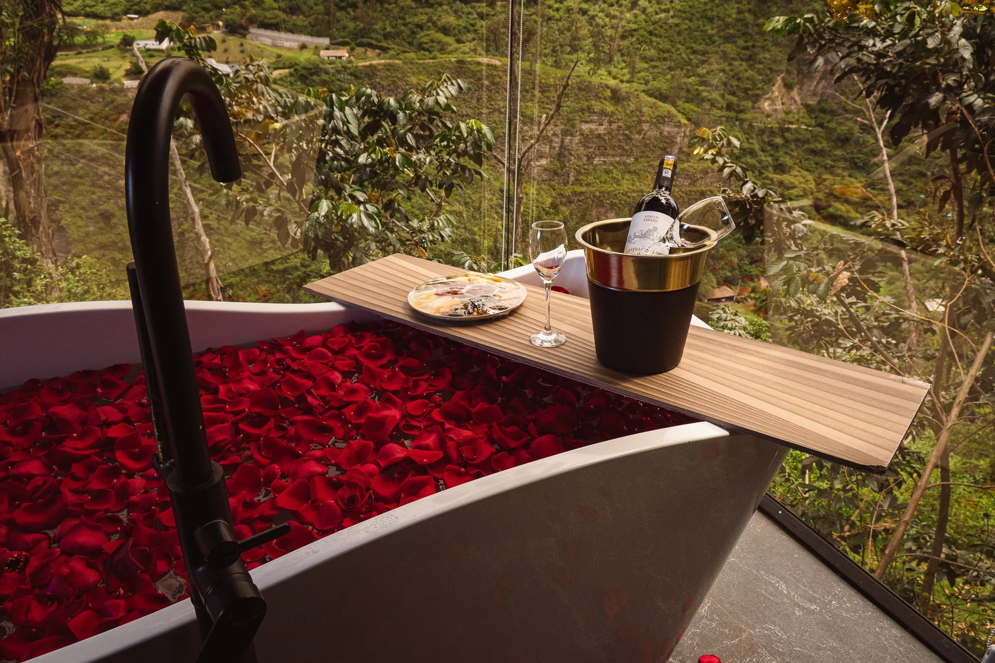 A bathroom with a bathtub filled with red rose petals, a wooden tray holding a plate of cheese, a glass of wine, and a bottle of wine inside a gold ice bucket. There is a black faucet, and a scenic view of green, hilly landscape with trees and houses through a large window.