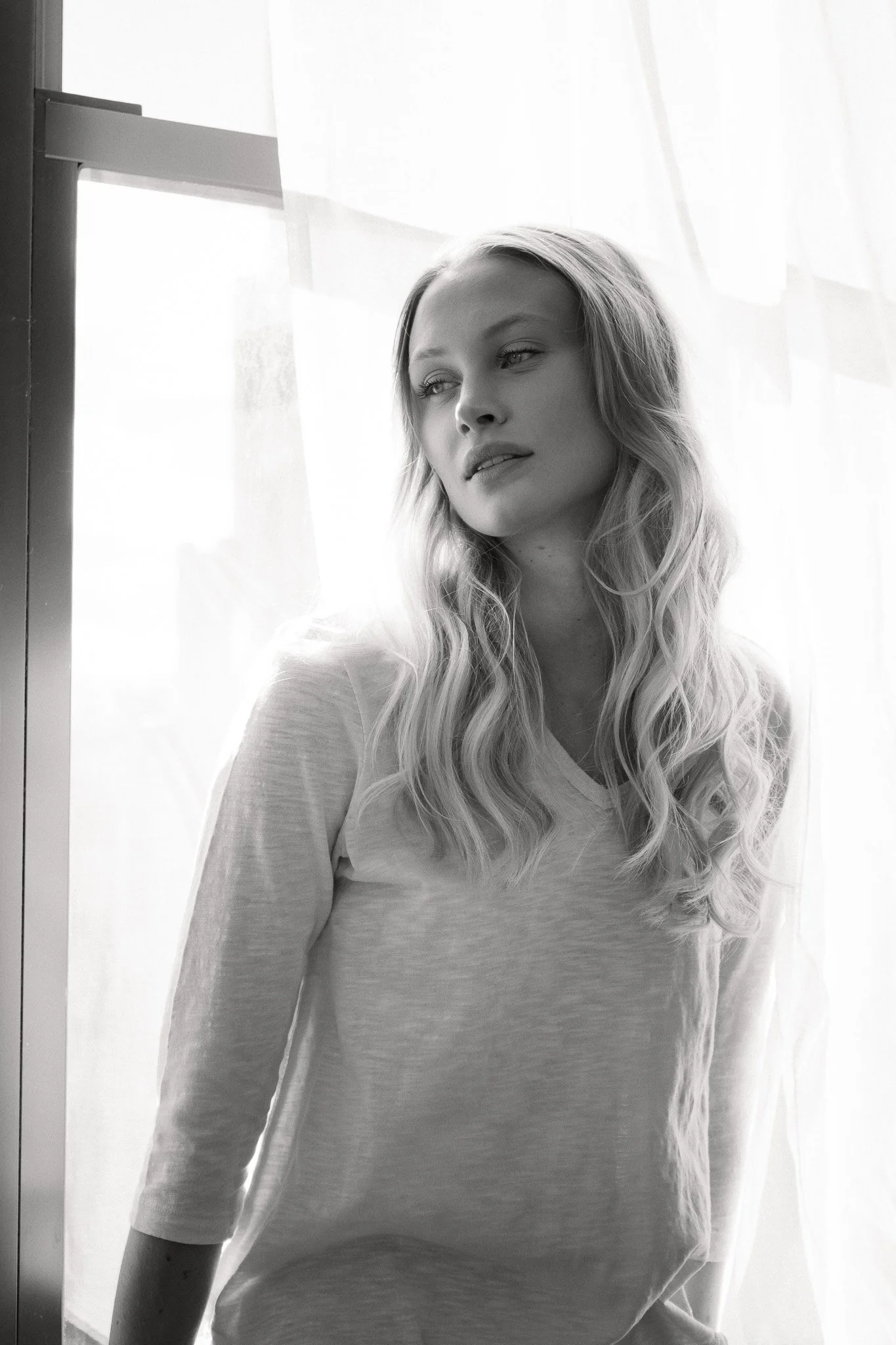 Black and white photo of a woman with wavy hair standing near a window with sheer curtains, looking to the side.
