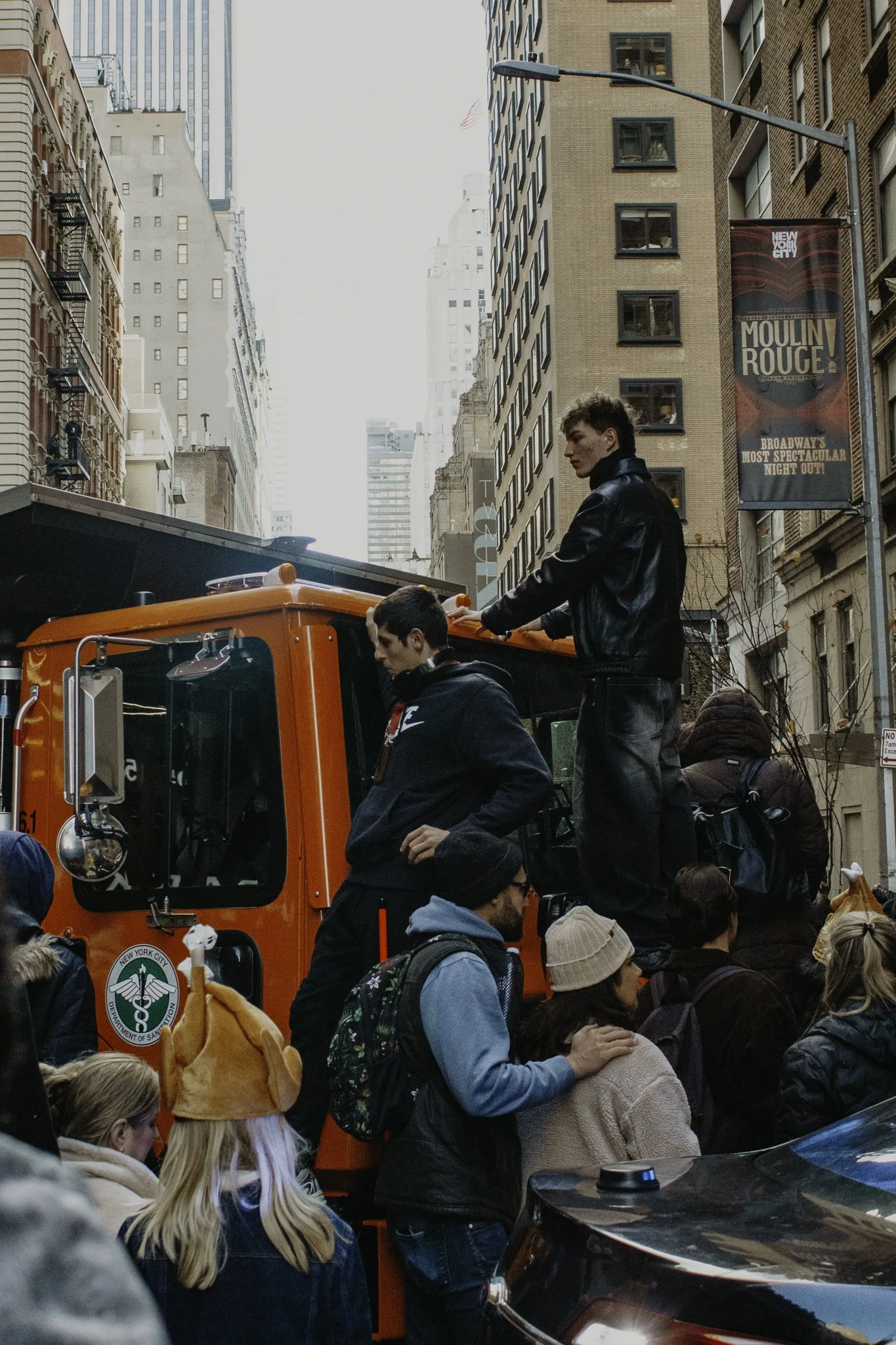 People gathered on a city street with tall buildings, with some standing on an orange fire truck. A man in a black leather jacket is on top of the fire truck, looking to the right. There are various individuals including a woman with blonde hair and 
