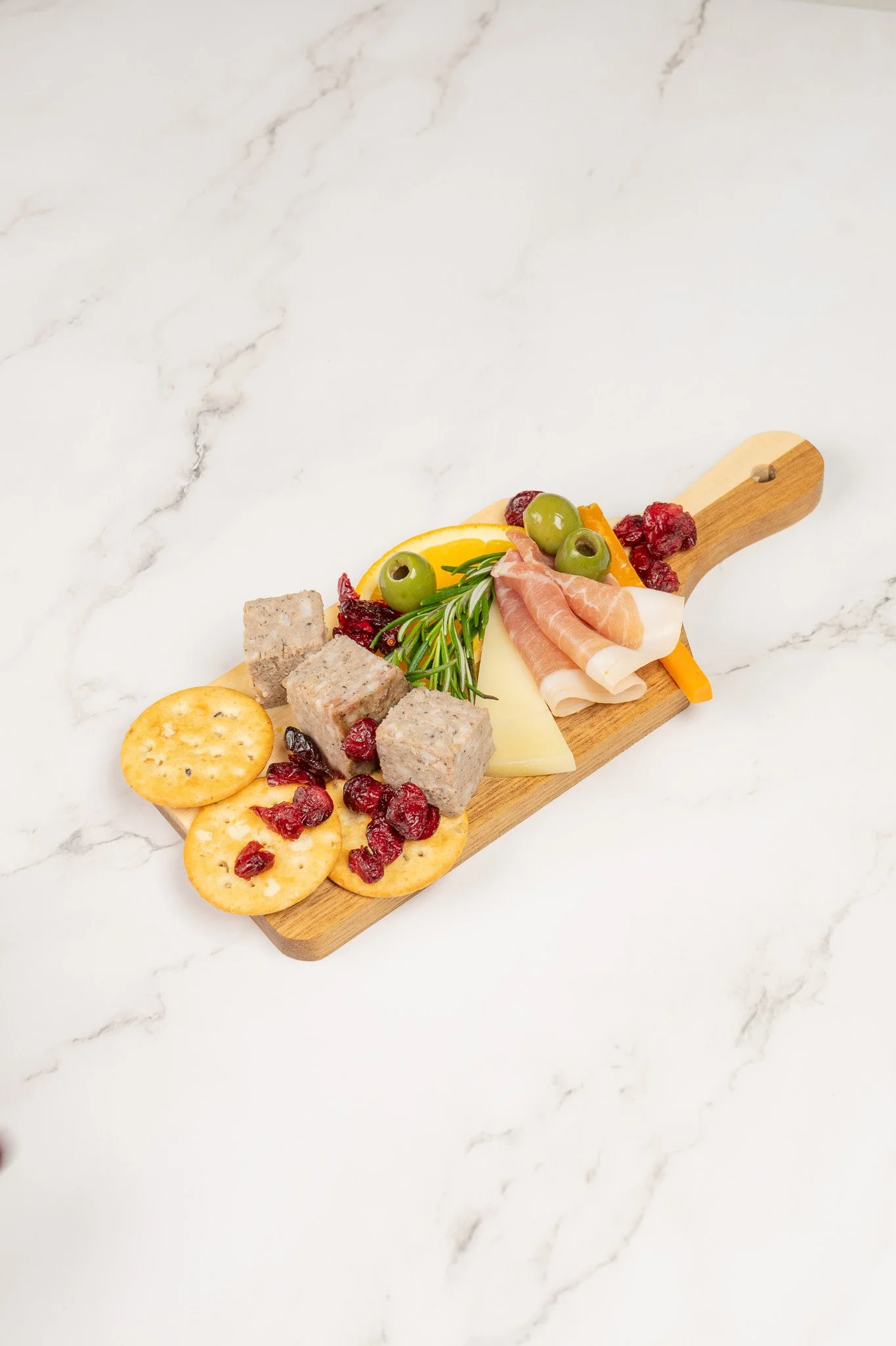 A wooden serving board with crackers, cubes of cheese, slices of cured meat, green olives, cherry tomatoes, cranberries, and a sprig of rosemary on a white marble surface.