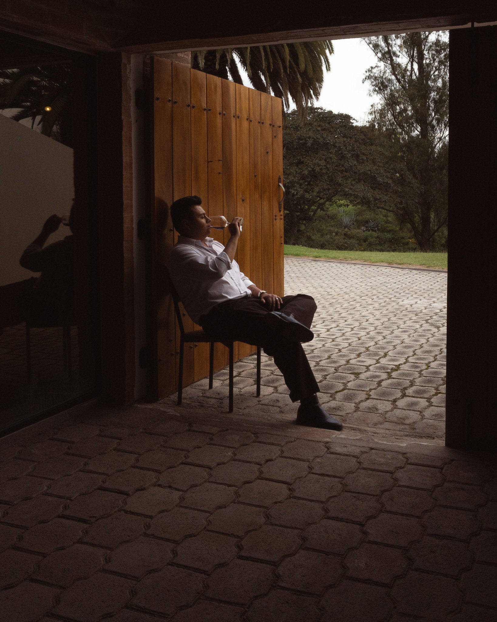 A man sitting on a chair outside near an open wooden door, drinking from a glass. The background shows trees and a paved area outside.