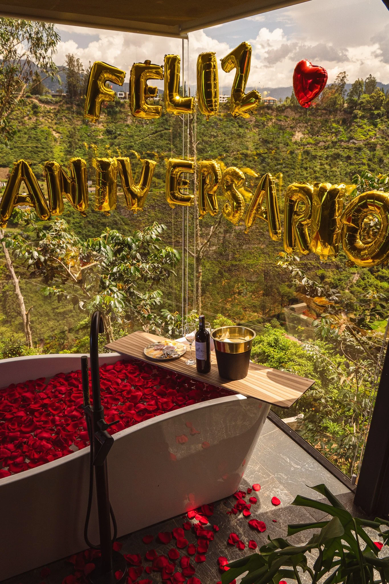 A bathtub filled with red rose petals, set in a scenic outdoor terrace with a view of a hilly area with green trees and houses; gold letter balloons spell out "FEEL" and "ANY" with a red heart-shaped balloon nearby. There is a table with a bottle of 