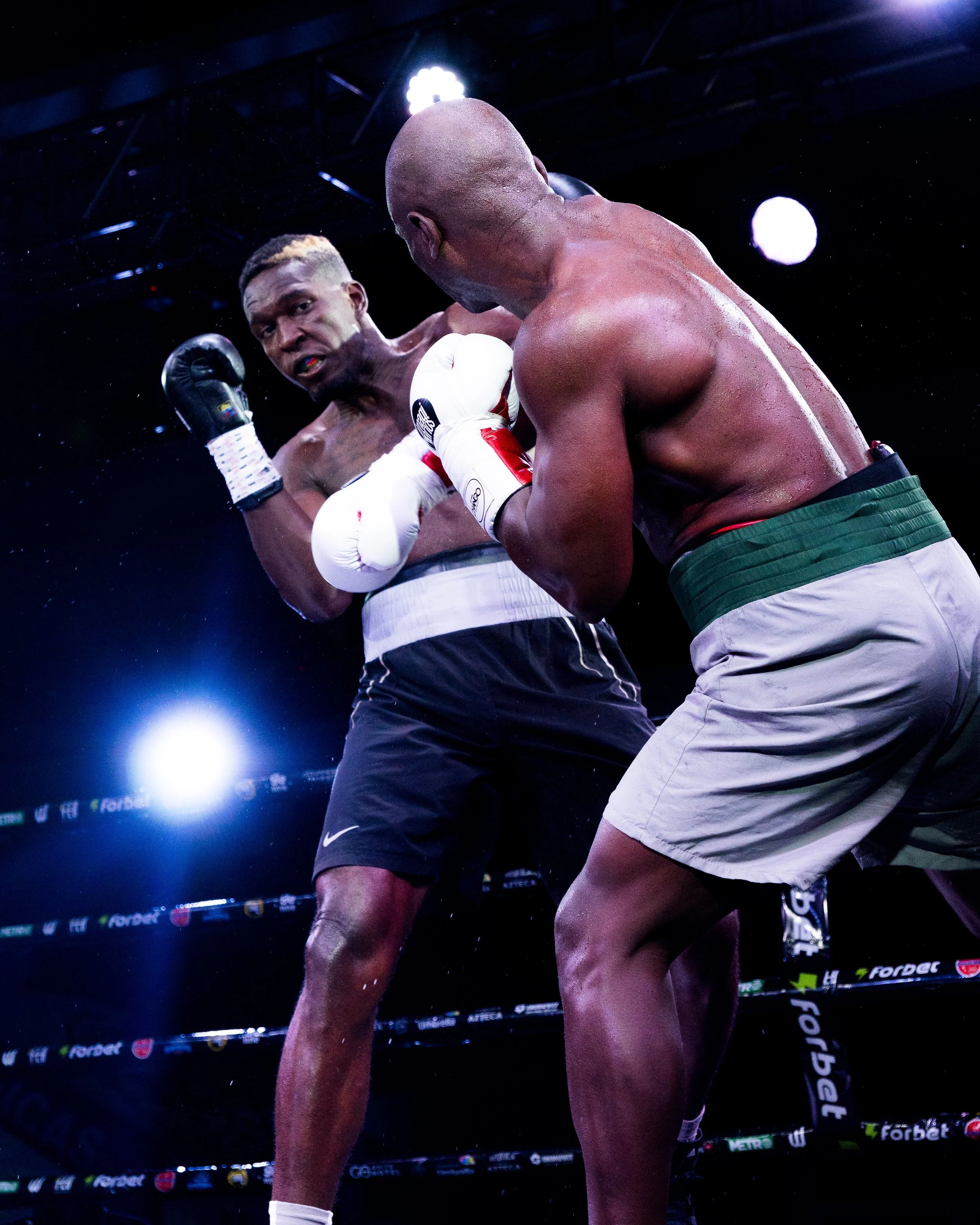 Two male boxers in a boxing ring during a match, one delivering a punch to the other's face, under bright overhead lights.