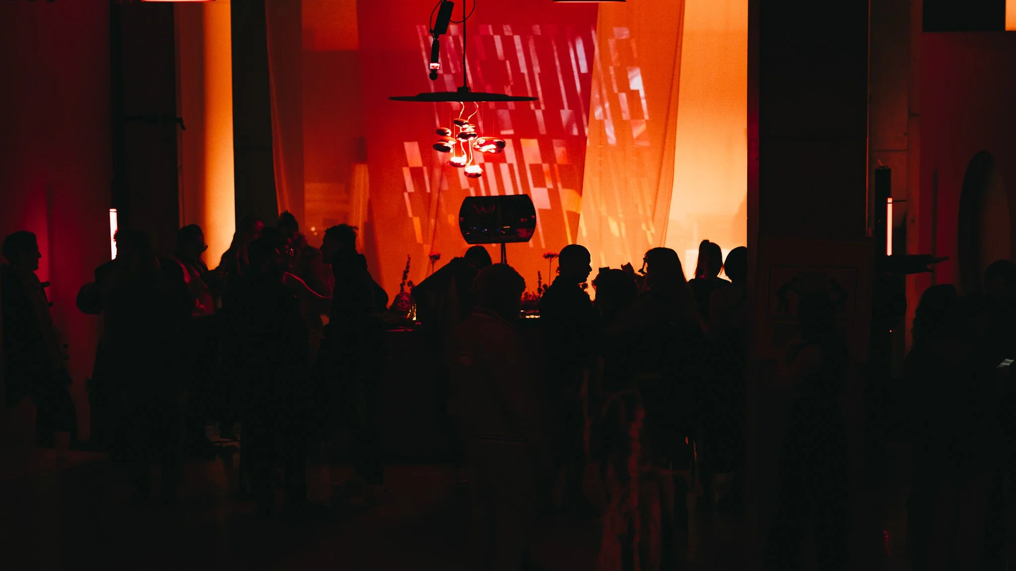 People dancing and socializing in a dimly lit venue with orange and red lighting, and a large abstract projection on the wall in the background.