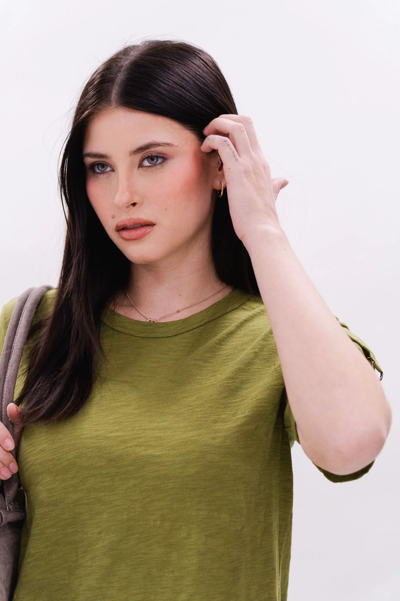 A woman with long dark hair, light skin, and blue eyes, wearing an olive green T-shirt, a gold necklace, gold hoop earrings, and holding her hair with one hand, standing against a white background.