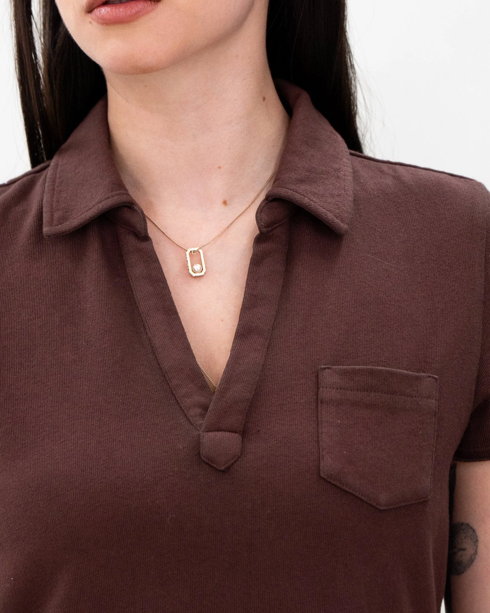 A close-up of a woman's neck and chest area, showing a necklace with a rectangular pendant containing a pearl, and part of her lower face and lips. She is wearing a collar shirt in a dark mauve color.