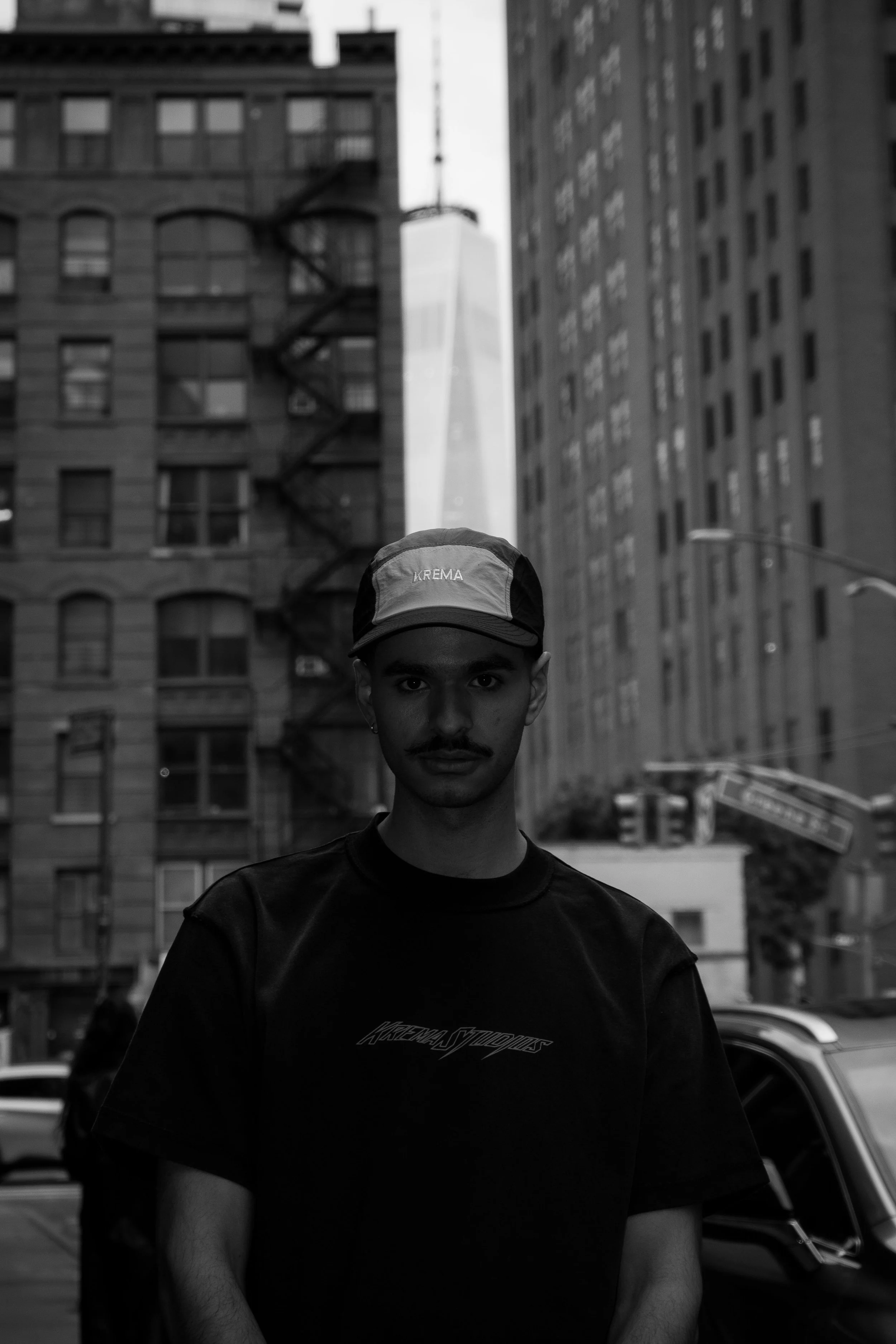 A young man with a cap and mustache standing on a city street in black and white, with tall buildings and skyscrapers in the background, including One World Trade Center.