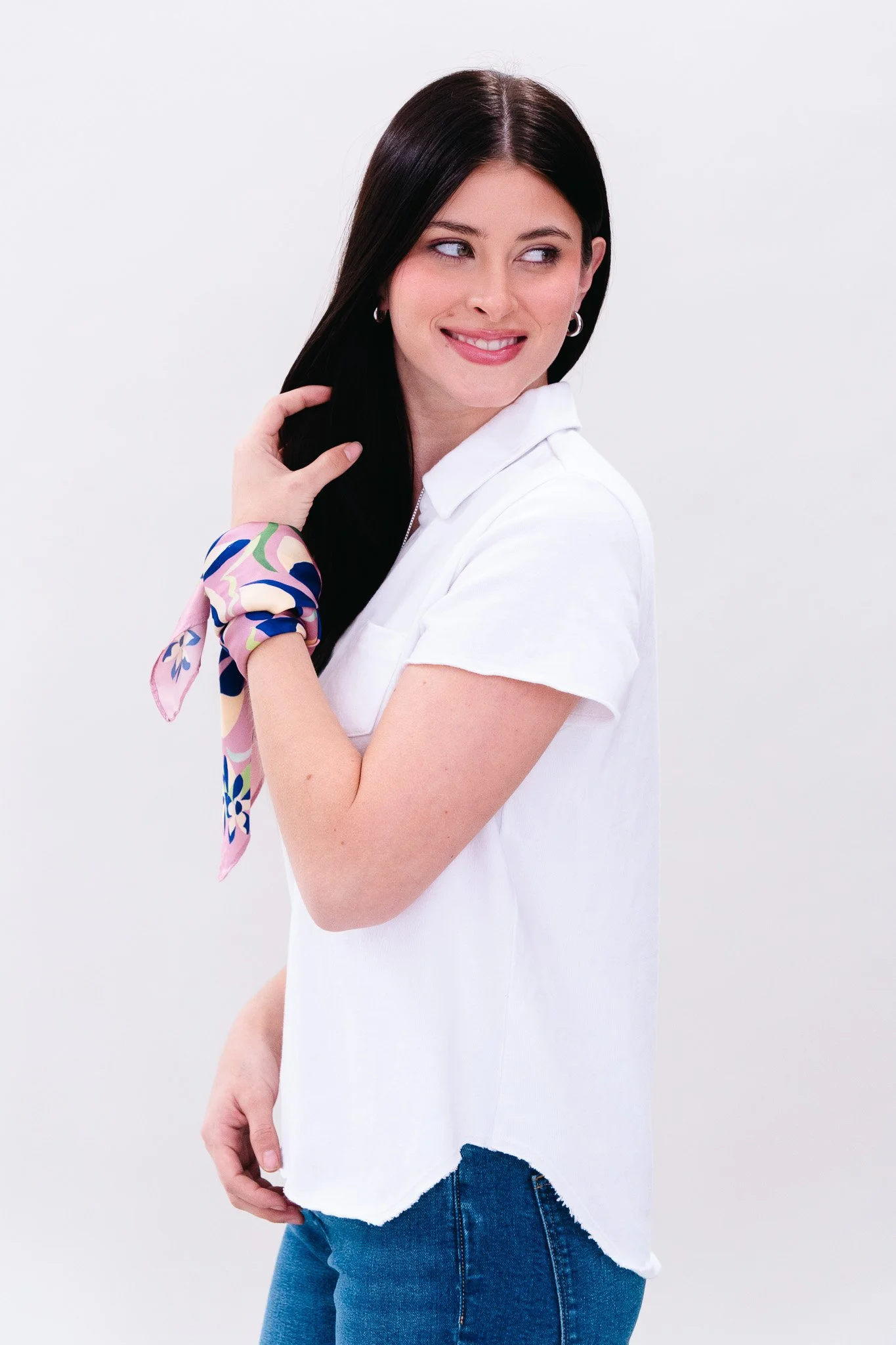 A woman with long dark hair smiling and looking to her left, wearing a white shirt, blue jeans, and a pink scarf with colorful patterns tied around her wrist, standing against a plain white background.