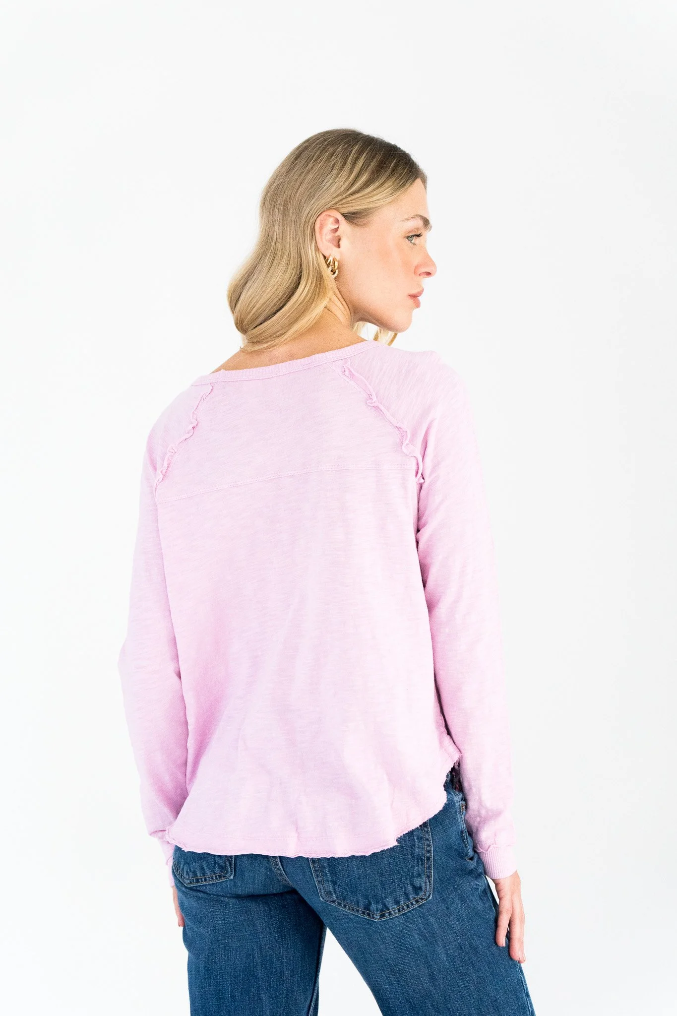 A woman with blonde hair, wearing a light pink long-sleeve shirt and blue jeans, standing against a white background, turned slightly to the right.