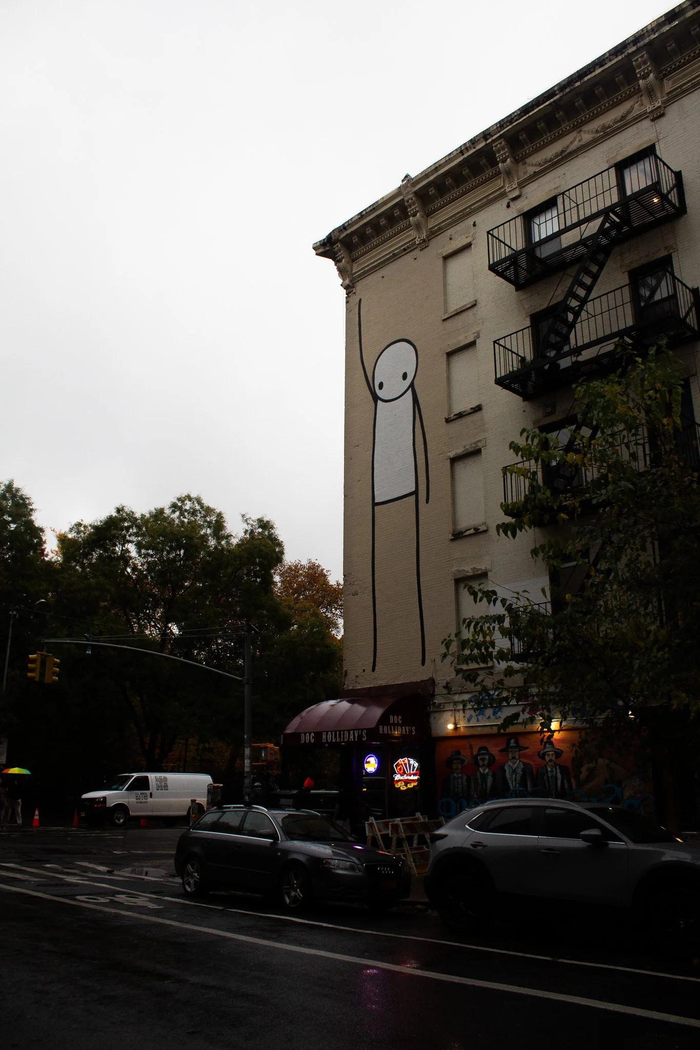 Street view of a building with a large minimalist mural of a tall, thin, smiling stick-figure person on the side wall. Nearby is a bar called 'Doc Holliday's' with neon lights and artwork of four men in hats on the lower wall. Cars parked in front an