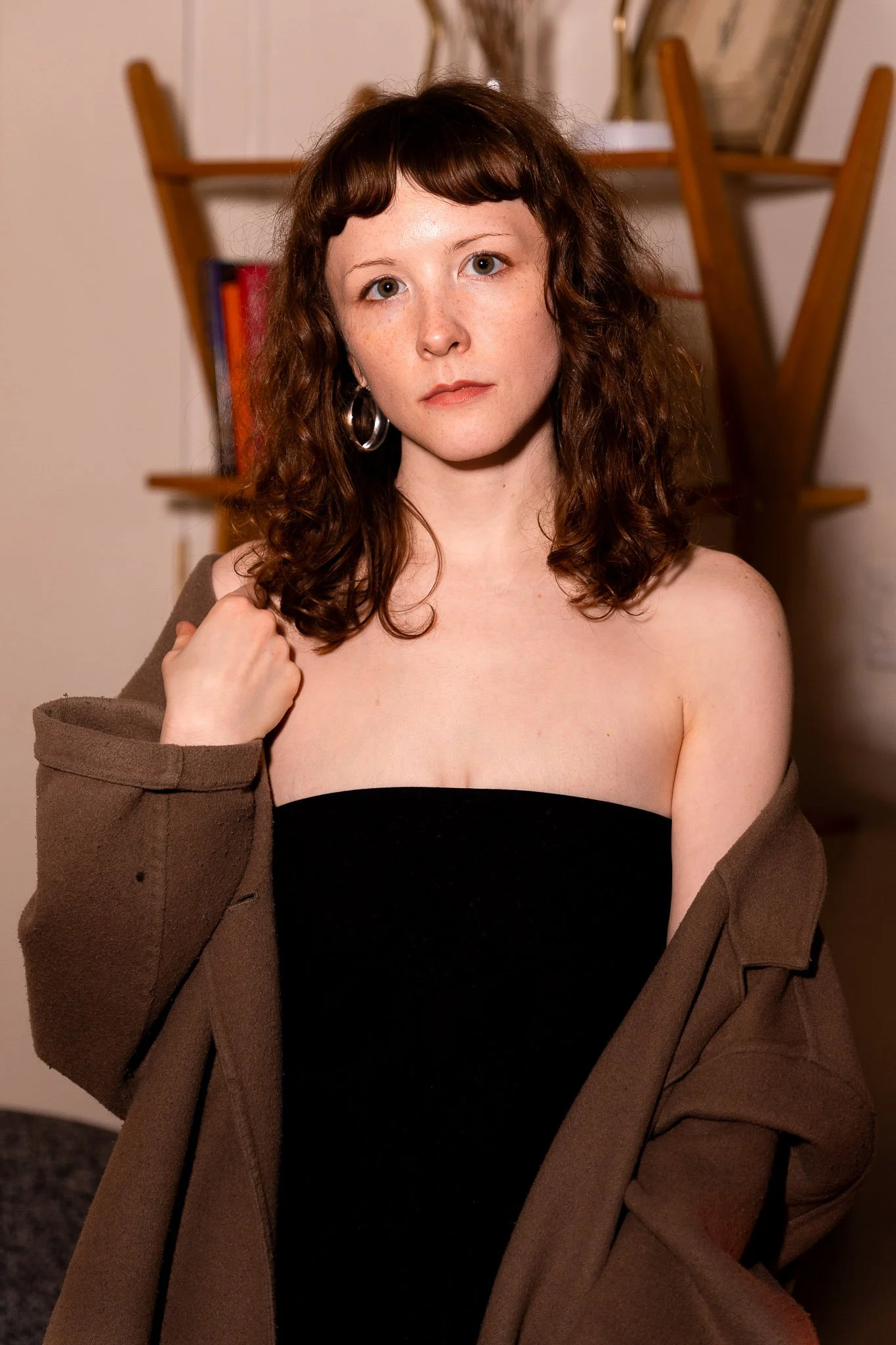 A woman with short curly brown hair and hoop earrings wearing a strapless black top and an open brown jacket, posing indoors with a blurred background of wooden furniture and framed artwork.