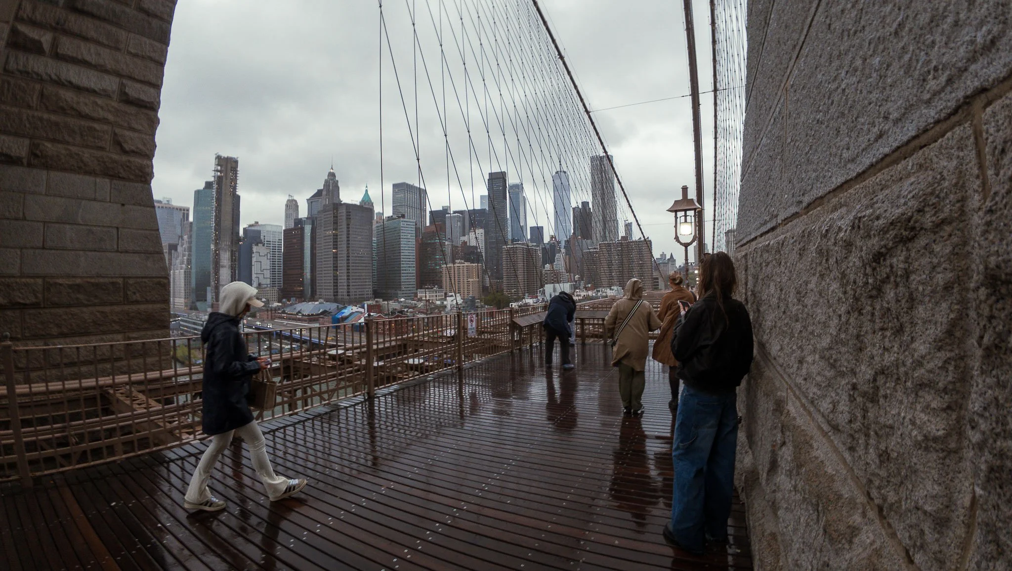 View of New York City skyline from the Brooklyn Bridge walkway on a rainy day, with several people walking and standing on the wet wooden platform, and the cityscape visible through the bridge's cables.