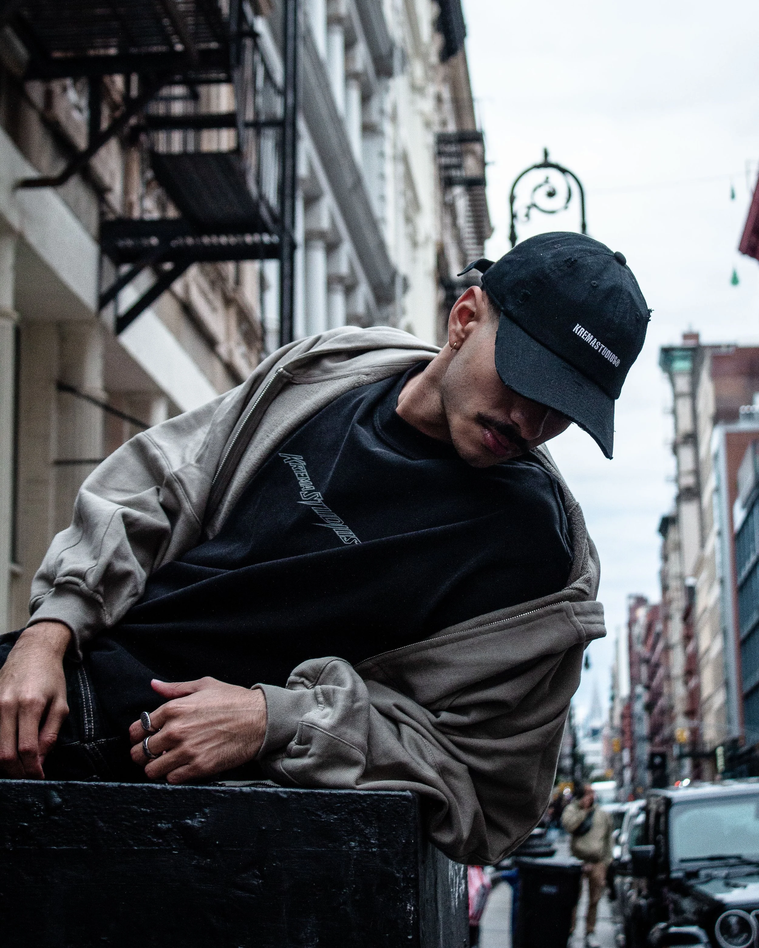 A person leaning on a black structure outdoors in an urban setting on a cloudy day, wearing a black cap, black shirt, and beige jacket.