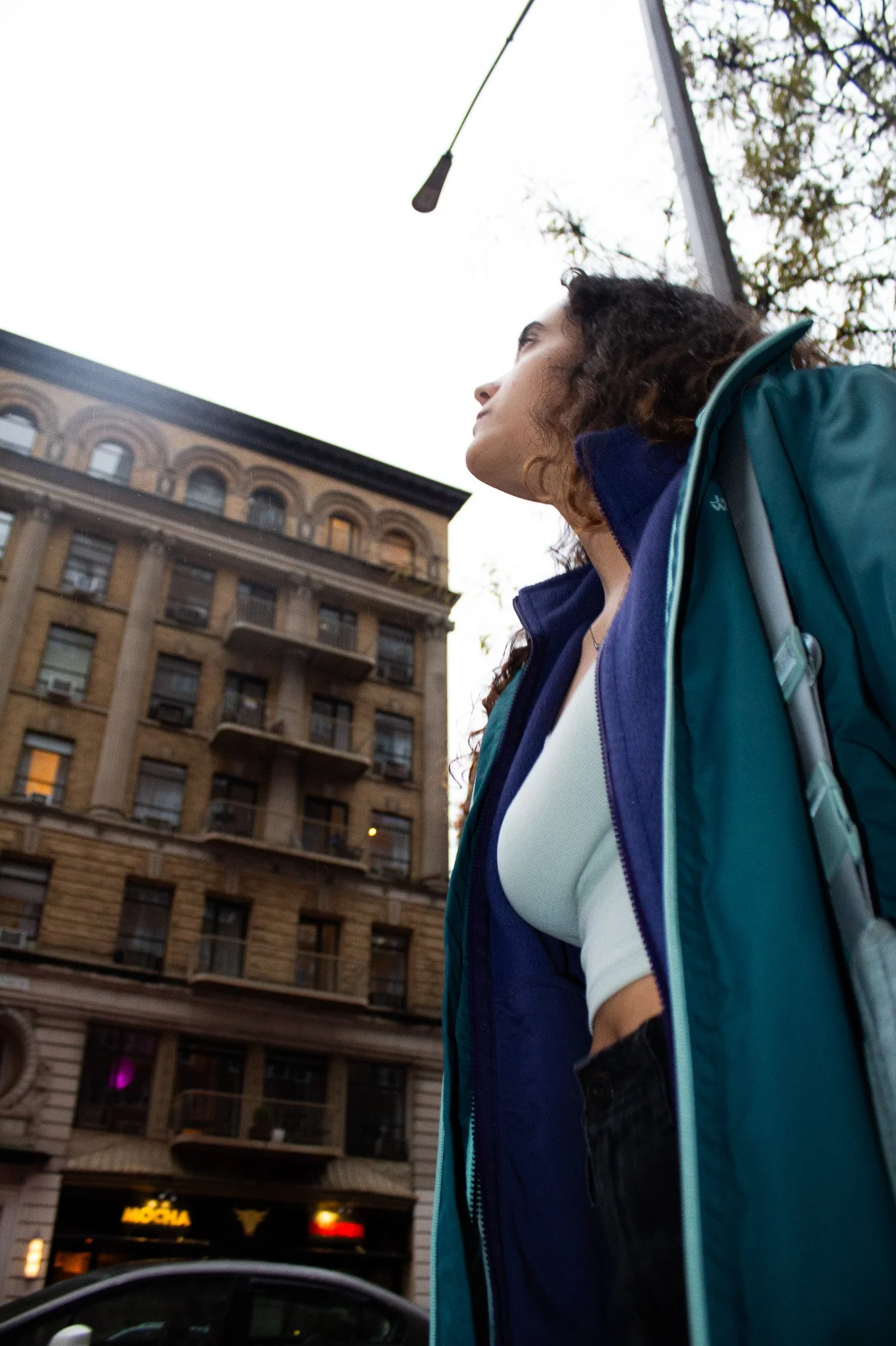 A woman with curly hair wearing a teal jacket and white top looking up at an overhead streetlight against a city building background.