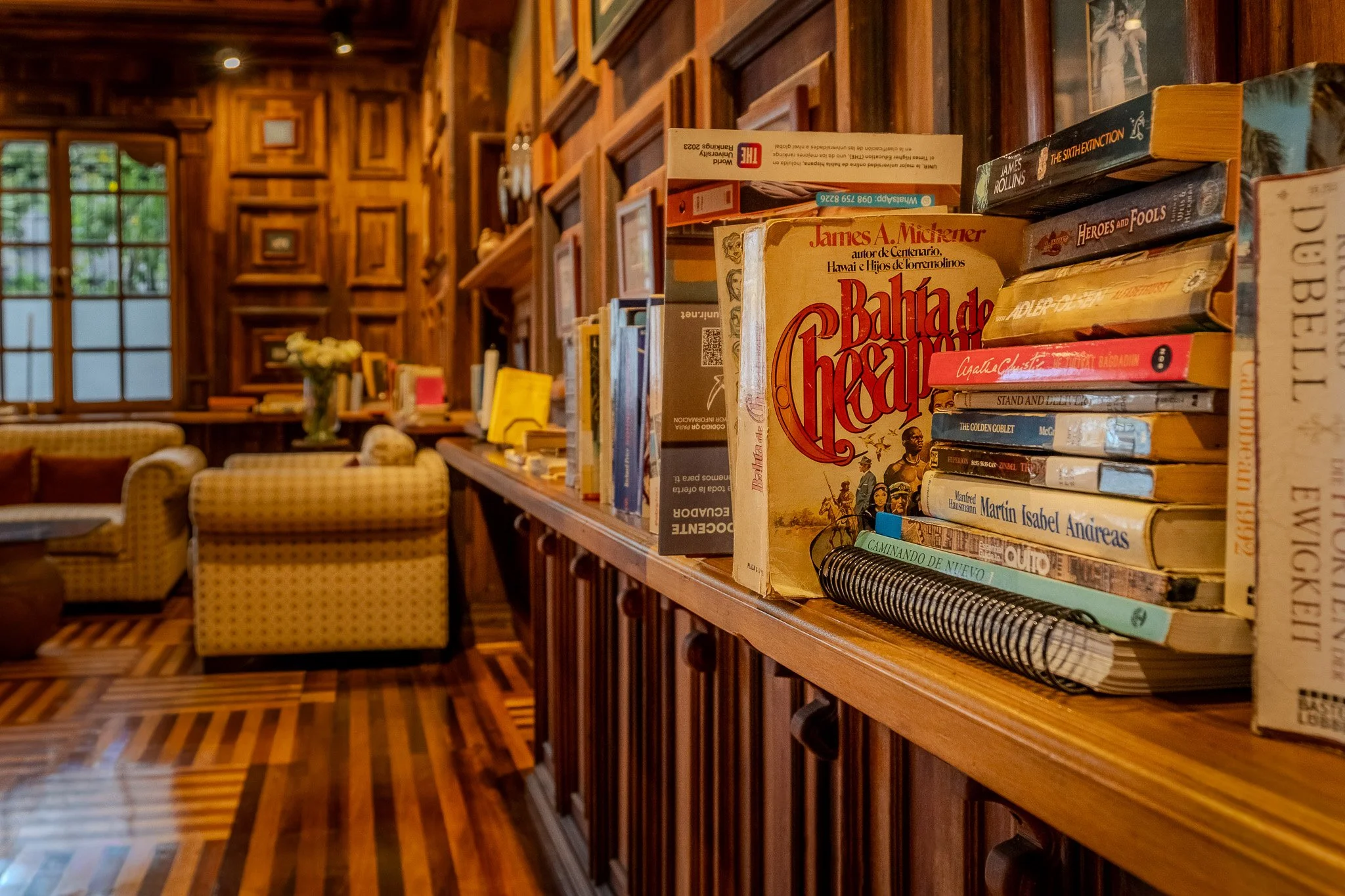 A bookshelf filled with books in a cozy, wood-paneled room with large windows and comfortable armchairs.