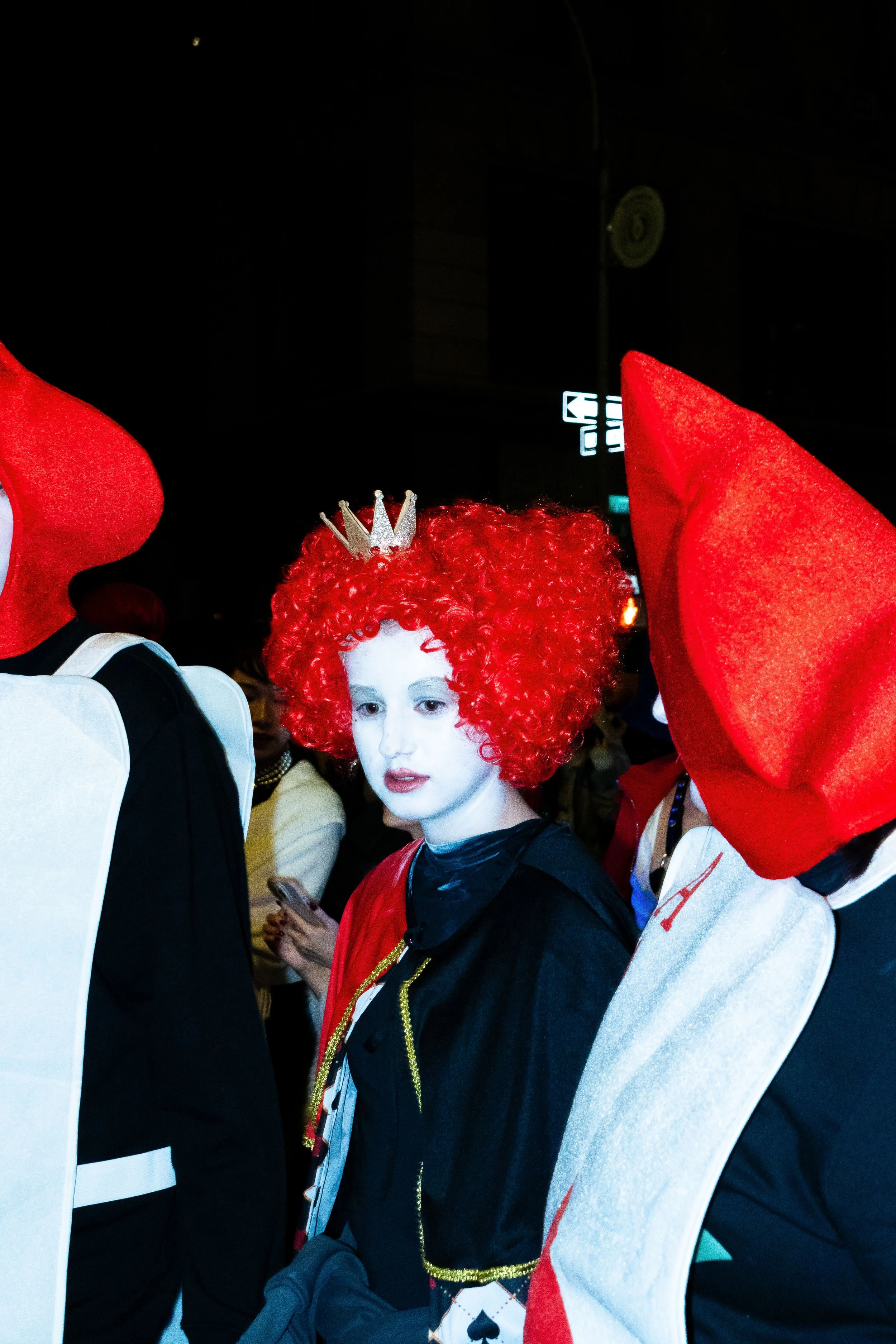 Person dressed as the Queen of Hearts from Alice in Wonderland, wearing a red, curly wig, a small crown, and white makeup, standing among others in costume at a nighttime event.