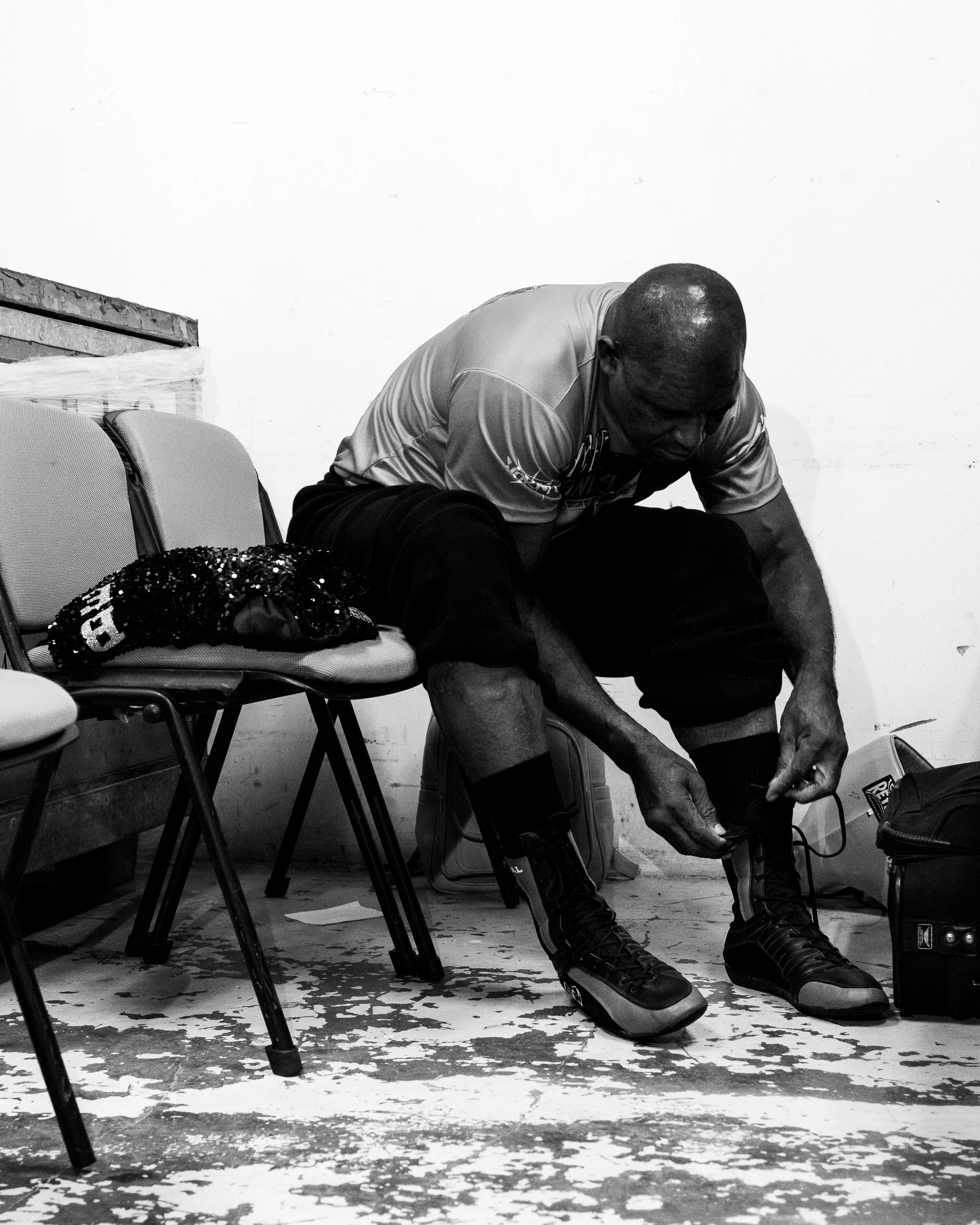 A man is sitting on a chair and tying his shoelaces, in a room with white walls. There are other chairs next to him, and various items including a backpack, a beaded bag, and some papers are visible on the floor and on the chairs.