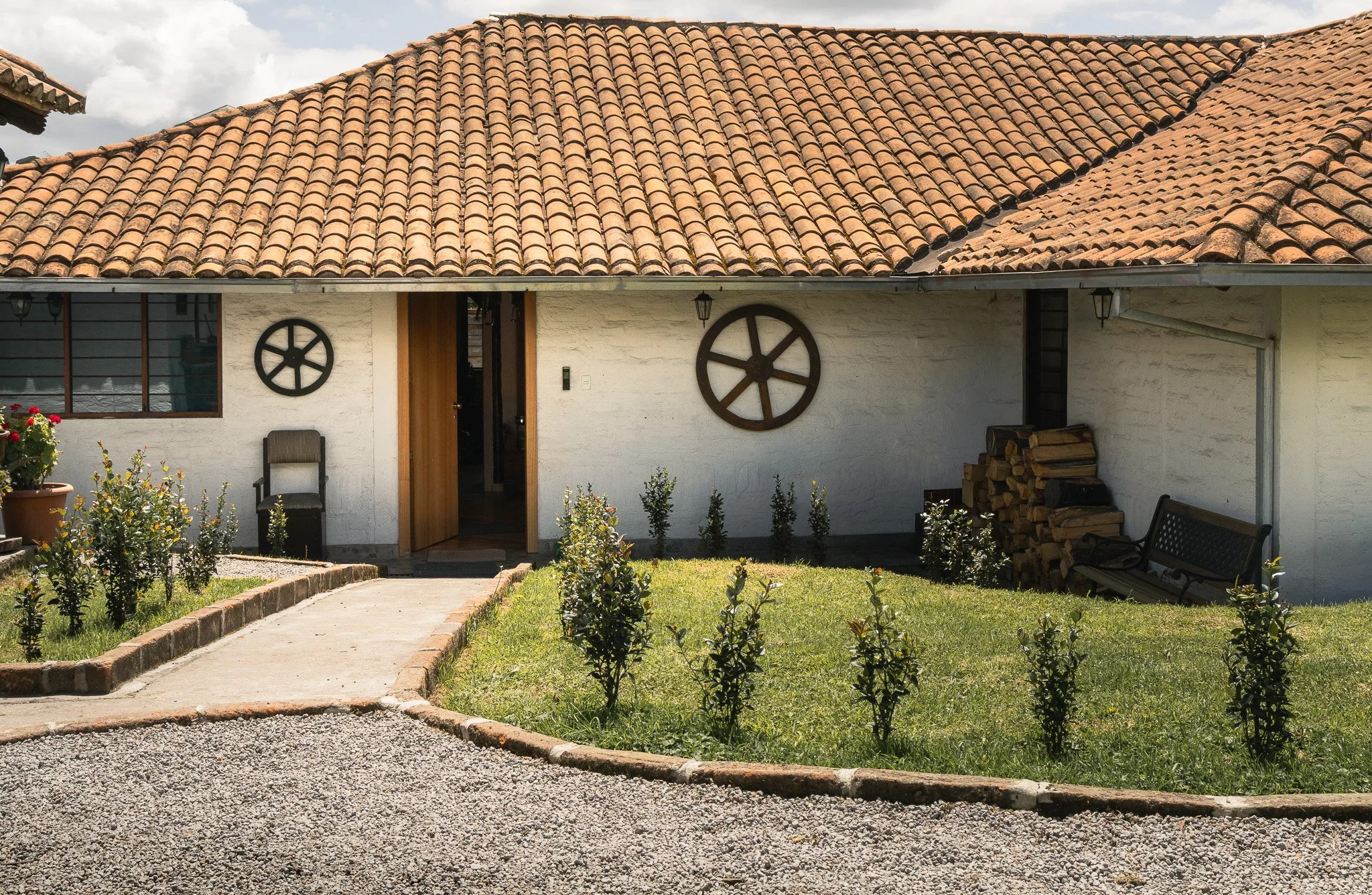 Front view of a house with a tile roof, white exterior walls, and decorative wagon wheels on the wall. A concrete pathway leads to the entrance, and there are plants and flowers in the garden area. Firewood stacked on the side of the house.