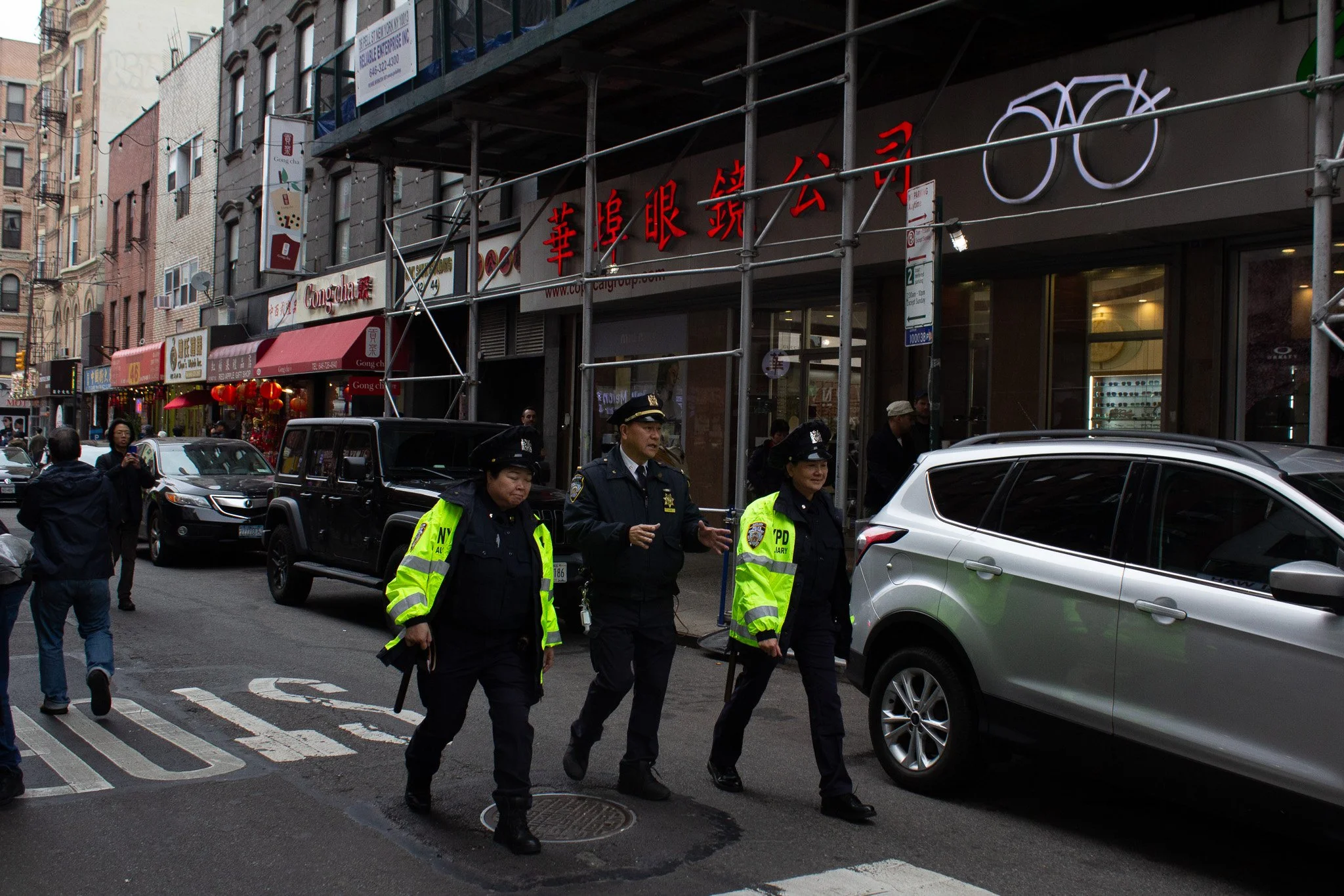 Three police officers walking on a city street in front of storefronts with Chinese signs; cars parked along the curb and pedestrians in the background.