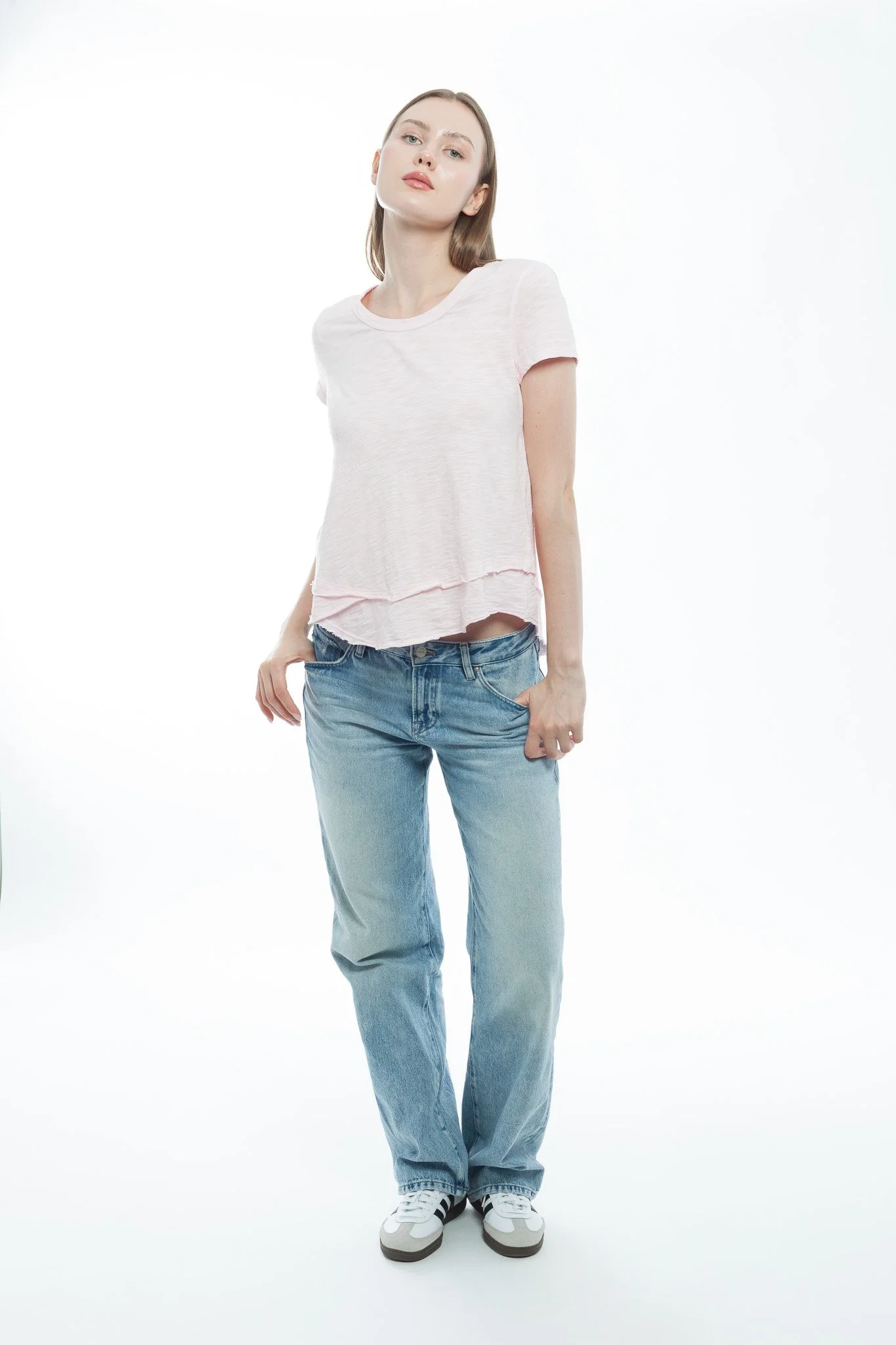 A young woman standing against a white background, wearing a light pink t-shirt, blue jeans, and white sneakers, with a confident expression.